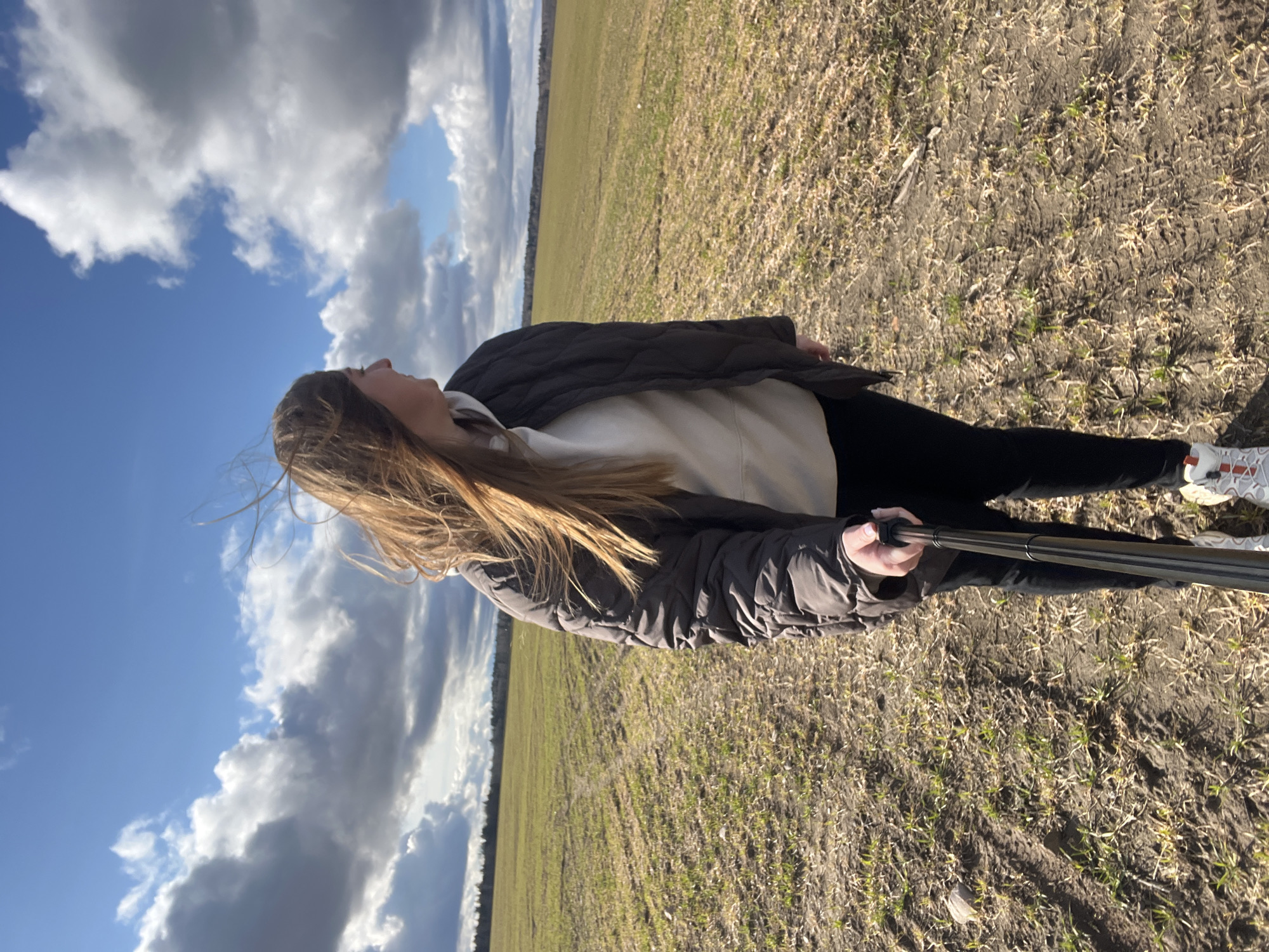 A woman with long brown hair is standing in a field with a blue and cloudy sky overhead. She is holding a selfie stick. She is wearing a white hoodie, black pants, a dark jacket, and white sneakers. The field is dry with sparse vegetation. The location is Ямуга, Russia.