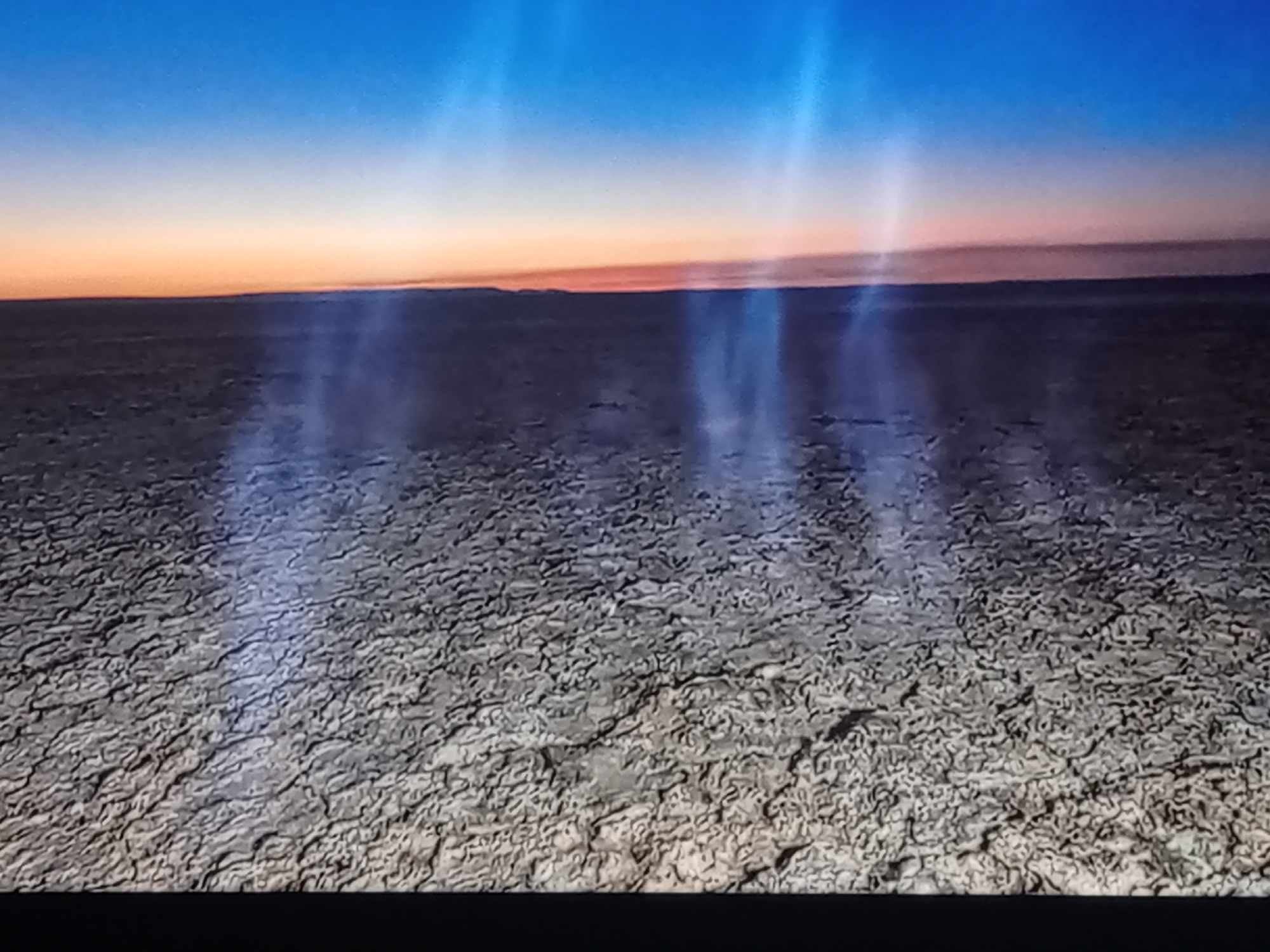 The image displays a vast, arid landscape under a gradient sky transitioning from deep blue at the top to orange and pink near the horizon. The ground is cracked and dry, resembling a salt flat or desert terrain. Several bright, ethereal shafts of light pierce downwards from the sky, illuminating patches of the ground and appearing as mist or fog. There are no visible people, structures, or distinct objects in the scene, suggesting a remote and desolate environment. The overall impression is one of natural phenomena occurring in an unpopulated area, possibly during sunrise or sunset.