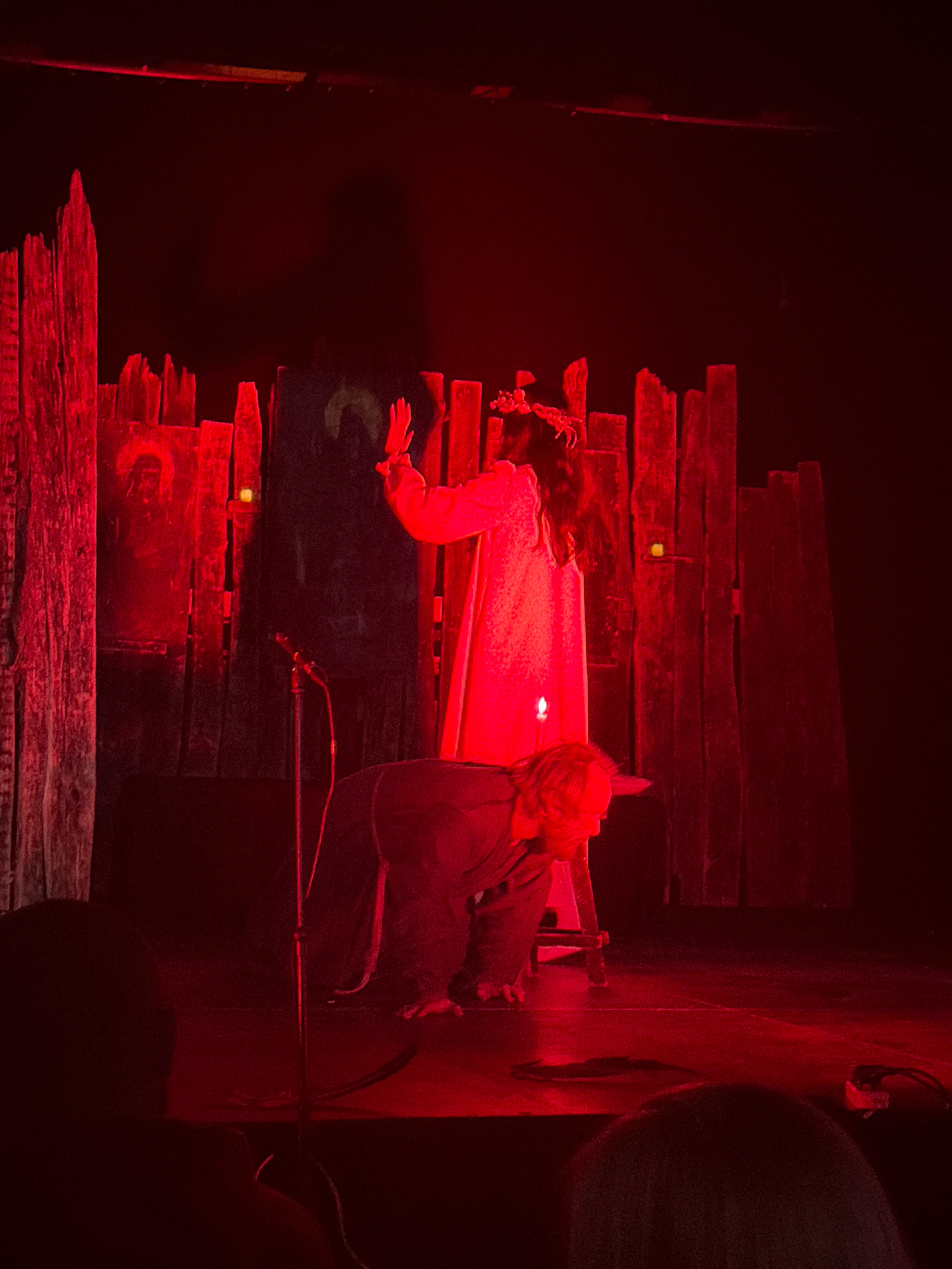This image captures a dimly lit stage, illuminated by red lighting, with two individuals. In the foreground, a person is crouched down, facing to the left. In the background, another individual stands, dressed in a long, light-colored garment and wearing a crown or headpiece. They are raising their left hand. Behind the standing figure, a wooden set piece resembling a fence or palisade is visible, with what appears to be a faint image of a face or figure on one of the wooden panels. A microphone on a stand is positioned in front of the wooden set. The overall atmosphere is theatrical and dramatic. The location is not discernable from the image itself.