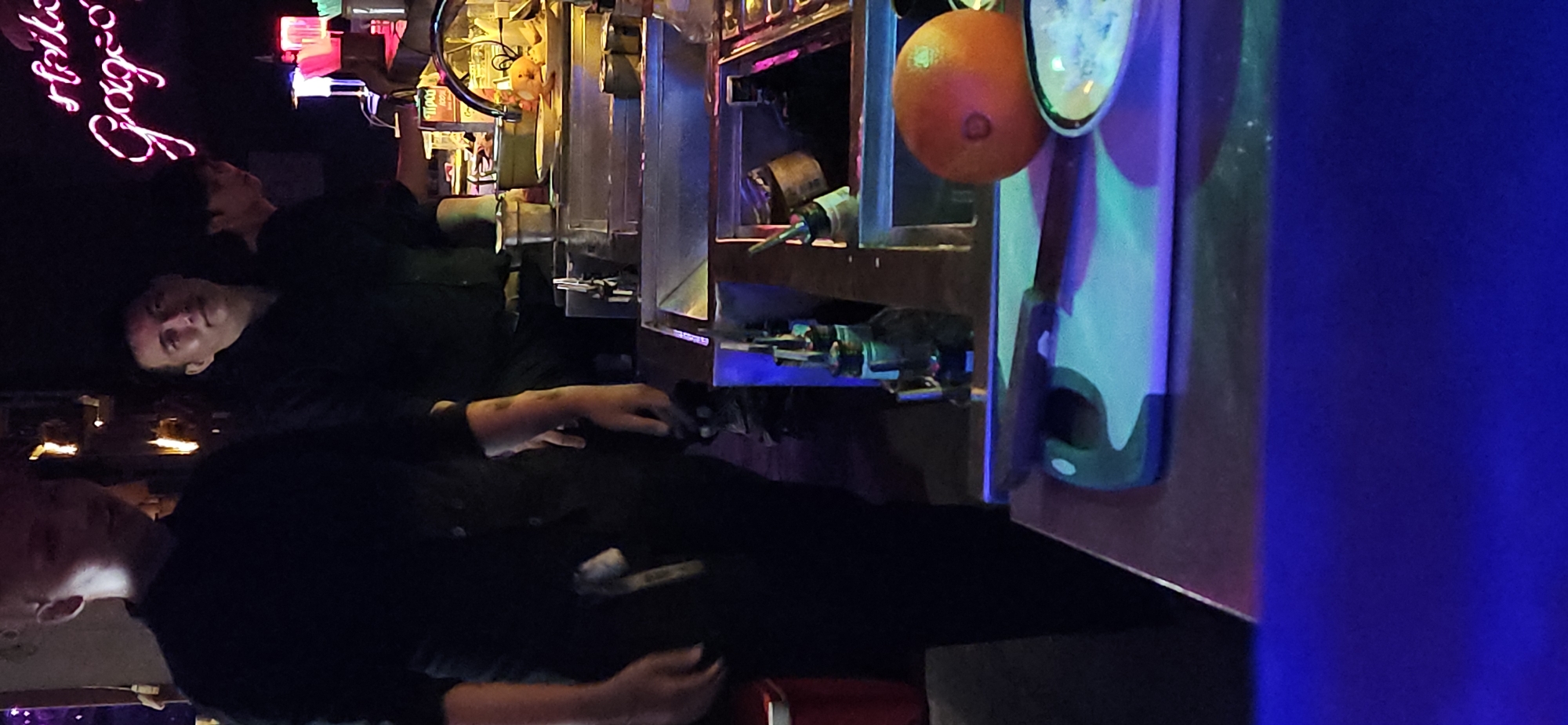 This image, presented with a vertical orientation, depicts a dimly lit bar scene at night. Two men are prominently visible, both wearing dark shirts. One man, likely a bartender, is positioned behind the counter, looking towards the viewer with a neutral expression. His hands are resting on the bar surface. Further back and to the left, another person's head is partially visible near a pink neon sign.
The setting is characterized by a long bar counter laden with bottles and bar equipment. An orange is conspicuously hanging near some bar tools. The lighting is low and atmospheric, with strong colored lights casting blue, green, and pink hues across the scene. A bright pink neon sign on the wall behind the bartender reads "Gorgeous" in a cursive font. The overall ambiance is typical of a bustling nightlife venue. The visible text confirms the presence of a bar or club, and the low light suggests a nocturnal setting.