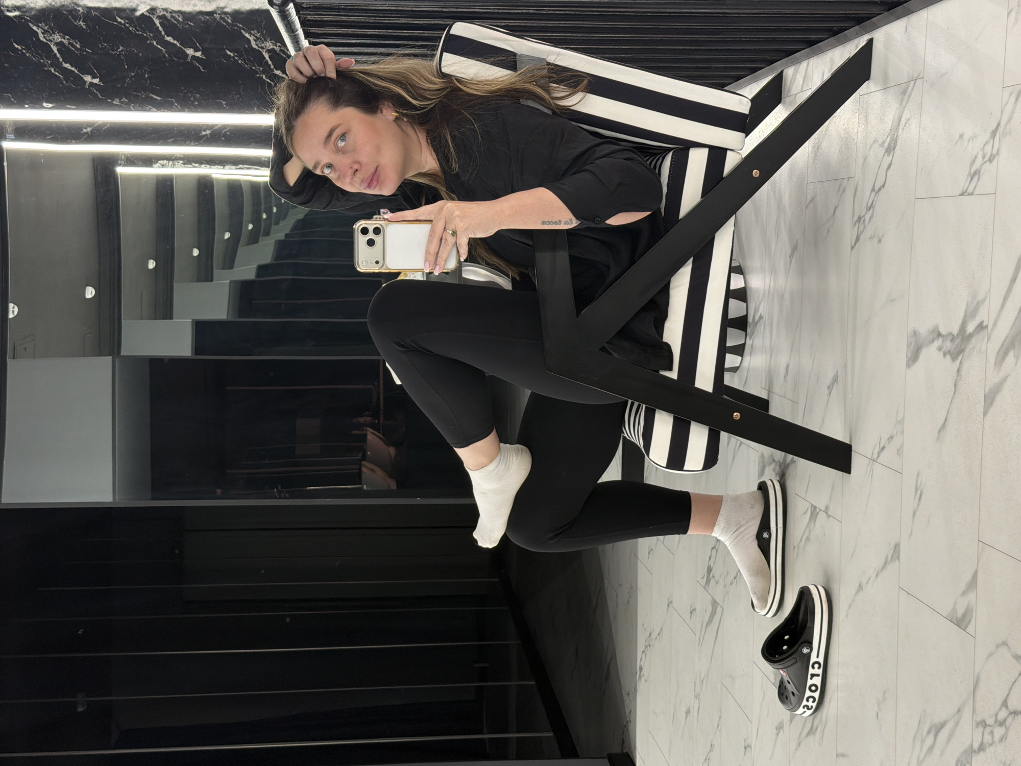 A woman takes a selfie in a modern, monochromatic interior, likely in Moscow, Russia. She is seated or leaning on a distinct black and white striped chair with a black frame. Her hair is light brown, and she wears a black long-sleeved top and black leggings. Her left arm, featuring a visible tattoo that reads "l'anima di...", rests on the chair's armrest. With her right hand, adorned with a ring, she holds a light-colored smartphone with a triple camera setup, pointing it towards a reflective surface. Her expression is neutral as she looks towards the camera's reflection.
Below her knees, her feet are clad in white socks. One foot rests on the light-colored, marble-patterned floor, next to which a black Crocs clog with "CROCS" visible in white lettering on its side lies unadorned.
The background features dark, highly reflective surfaces, possibly mirrors or polished walls, which reflect strip lighting and create an illusion of depth, revealing what appears to be a modern staircase or architectural elements within the space. The overall ambiance is contemporary and chic, dominated by black and white tones. The scene is brightly lit by artificial light sources.