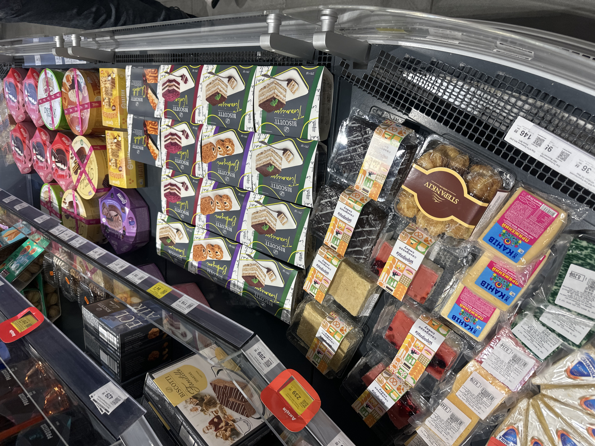 The image depicts a grocery store aisle in Ternopil, Ukraine, showcasing a variety of packaged baked goods and cheeses on display in refrigerated shelves. Prominently featured are boxes of "Biscotti" in Tiramisu and Bombonieri flavors, alongside other assorted cookie and cake boxes, some with decorative ribbon accents. Various packages of cheese are visible in the lower section of the display, including wedges and blocks with labels indicating brands and flavors. A red "Акція" (Sale) sign with a price of 239 UAH is attached to the shelf. The overall environment suggests a well-stocked supermarket with typical retail shelving and product arrangement. No individuals are clearly visible in the frame.