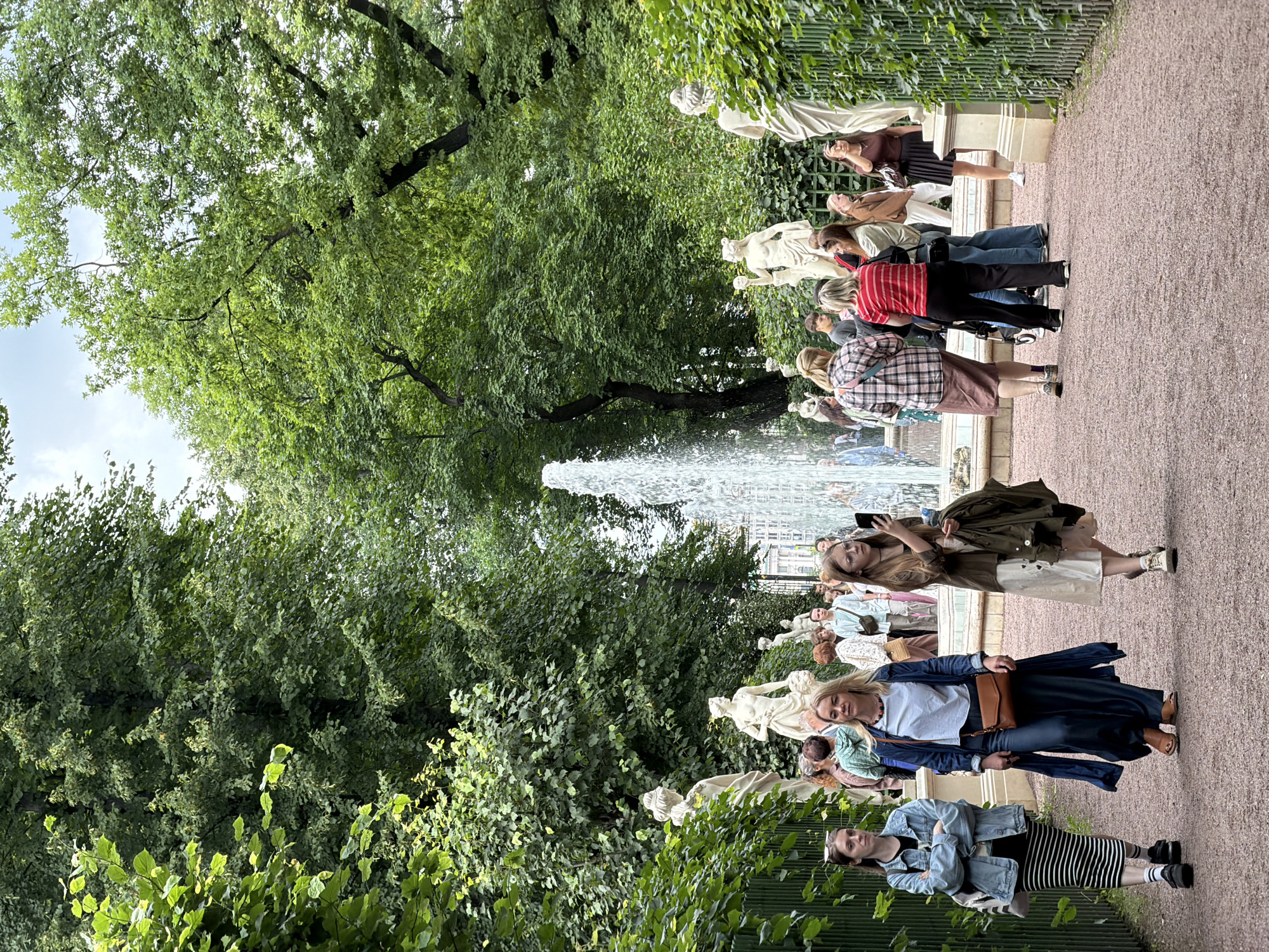 The image depicts a view of the Peterhof Palace gardens in Saint Petersburg, Russia, on a cloudy day. A tall fountain is visible in the background, with its water arcing upwards. Several people are walking along a gravel path lined with lush green hedges and marble statues. In the foreground, three women are walking towards the viewer. The woman on the left wears a denim jacket over a striped dress, the woman in the middle has on a long denim duster coat over a light blue top and wide-leg pants, and the woman on the right is in a light-colored dress and a khaki jacket. Other individuals, a mix of adults and children, are dispersed throughout the scene, some looking at their phones, others conversing, and some appearing to be admiring the statues. The overall impression is of a pleasant day spent in a well-maintained park with classical architecture and natural beauty.