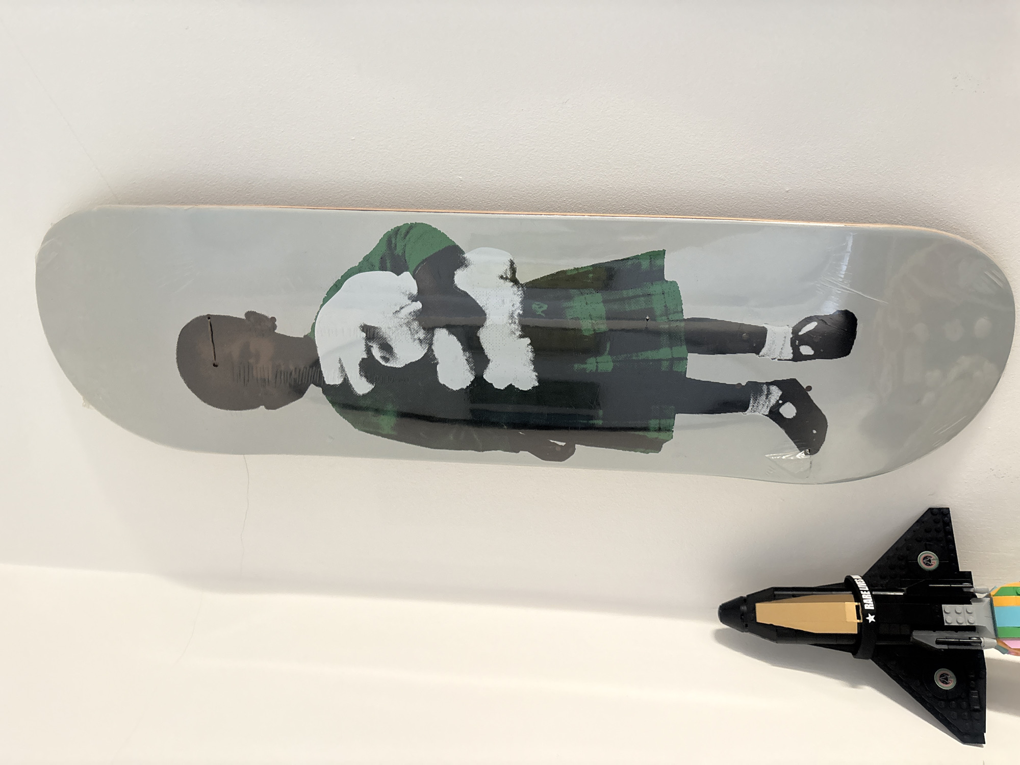 The image displays a skateboard and a LEGO model spaceship arranged on a white surface, likely a shelf or wall, against a plain white background.
The main subject is a skateboard, positioned horizontally and slightly angled. Its surface features an artistic depiction of a person, appearing to be a child or young individual, with dark skin, lying down or reclining. The figure wears a dark green and black plaid kilt or skirt, a matching green top, and black shoes. White, possibly furry or feathery, elements are visible on the arms and around the chest area. The entire skateboard is encased in clear protective plastic, indicating it is new or preserved.
Below the skateboard, to the right, is a black LEGO model spaceship. It has a pointed nose cone, a tan-colored section on its upper body, and broad wings. The rear of the spaceship features colorful thruster elements, including yellow, green, blue, and pink bricks. A white band around the middle section of the spaceship displays white text, partially visible as "RARE" and a star symbol. The remaining part of the text is obscured but appears to be part of a word like "DREAMS".
The setting suggests an indoor display, possibly in a home or gallery in London, United Kingdom. The lighting is even, with no harsh shadows, illuminating the white surfaces and the objects clearly. No activity is taking place; the items are stationary, presented as a collection or decorative pieces.