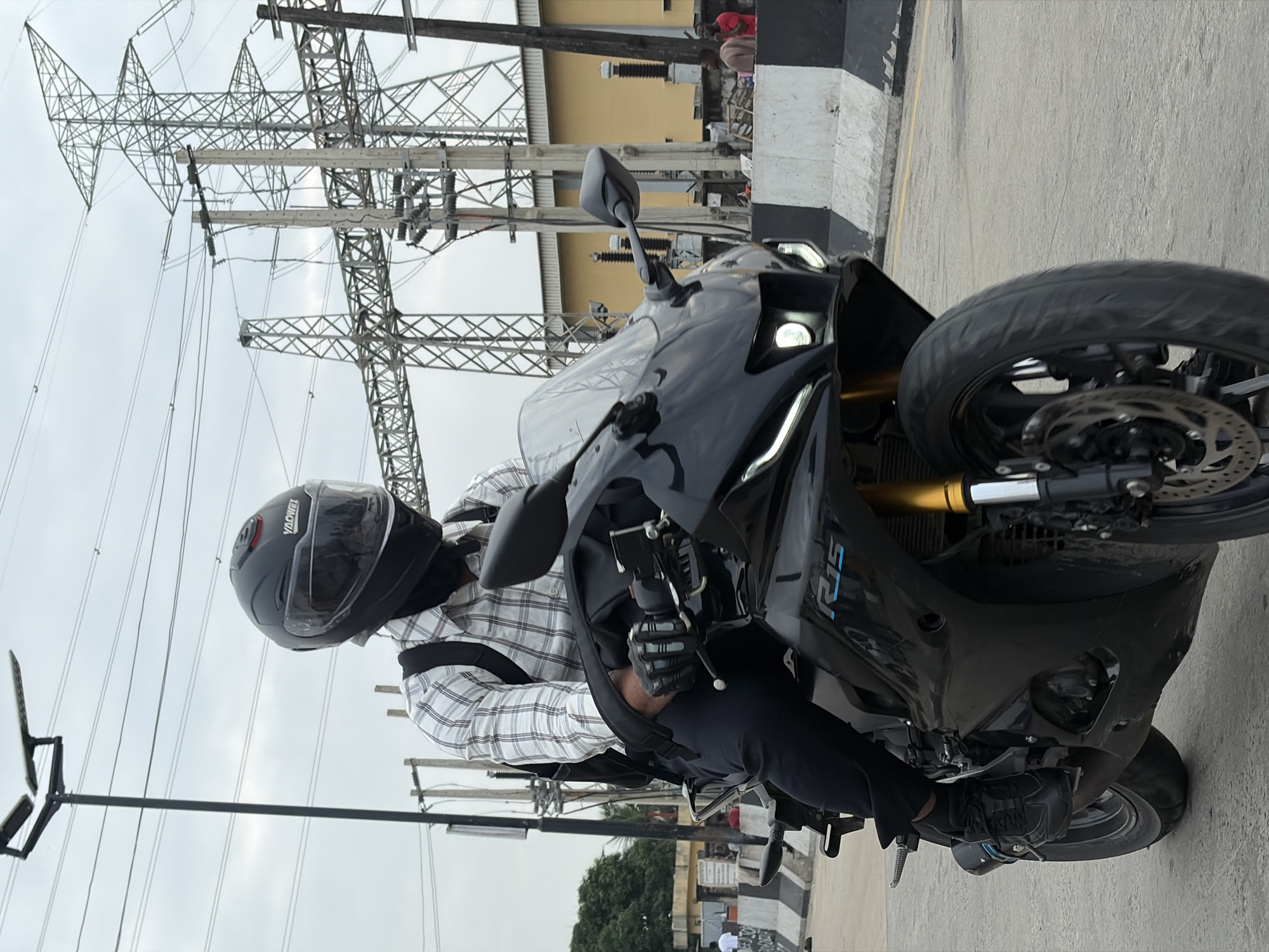 A person wearing a black helmet and a checkered shirt rides a black motorcycle on a road in Igboefon, Nigeria. The rider is wearing black pants and black shoes, and black gloves on their hands. The motorcycle has a blue "R15" logo on its side. In the background, there are tall metal towers and power lines against a cloudy sky. A yellow building is visible behind the power lines, and a concrete barrier with black and white markings lines the side of the road. A street light with solar panels is also present to the left of the frame.