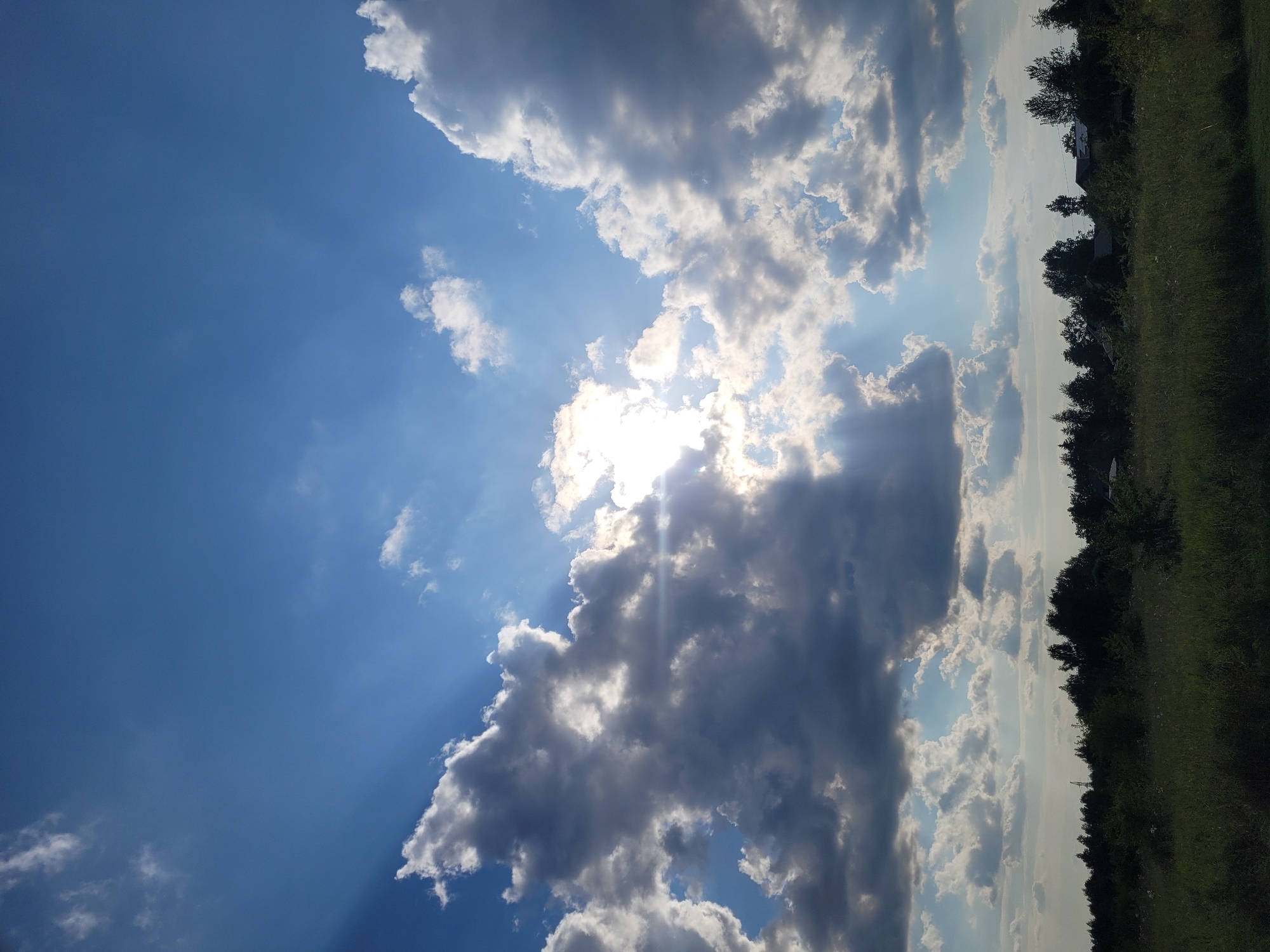 The image depicts a vibrant sky with a mixture of white and dark gray clouds, illuminated by bright sunlight streaming through. The sun is partially obscured by a large, dark cloud formation on the left side of the frame, casting dramatic rays of light across the sky. The lower portion of the image shows a dark green treeline against a lighter green grassy field. A few indistinct structures, possibly houses, are visible nestled amongst the trees on the right side of the frame. The overall impression is that of a dynamic and somewhat moody sky over a rural or semi-rural landscape. The specific location in Jūrmala, Latvia, cannot be determined from the visual information alone.