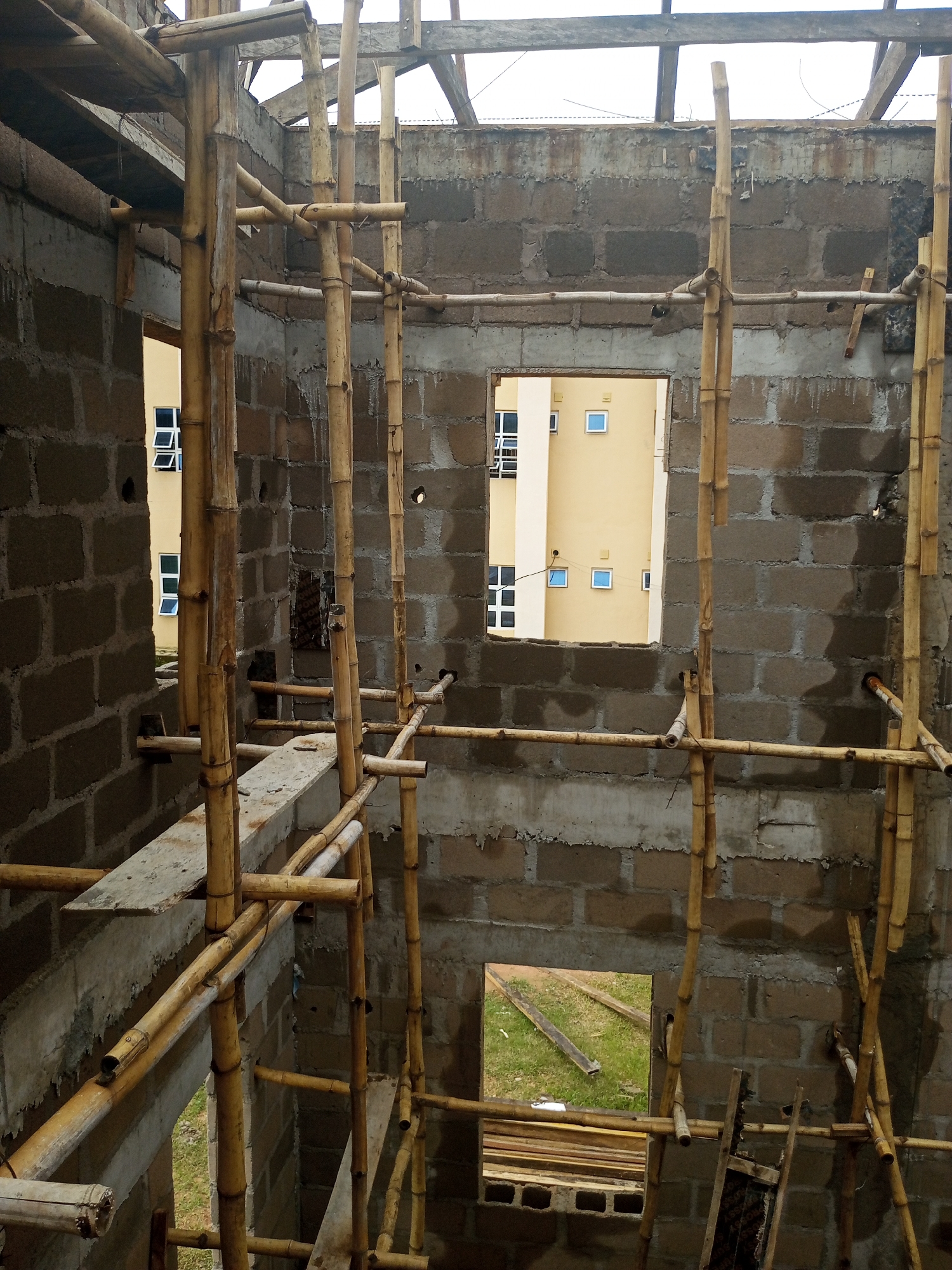 This is a medium-angle shot of a building under construction, viewed from an interior space looking outwards and upwards. The construction appears to be in an early stage, with concrete block walls that are partially plastered. Bamboo scaffolding is extensively used throughout the scene, forming a complex network of poles and supports within and around the structure.
The building has several openings, including window frames and a larger aperture that reveals another building in the background. This external building is painted a pale yellow and has numerous windows with white frames and blue panes, suggesting it is a finished structure. The sky above is overcast, indicating a cloudy day.
The foreground is filled with the raw construction materials and the bamboo scaffolding. Wooden planks are visible on the scaffolding, and some are also seen lying on the ground outside through an opening. The ground outside appears to be covered in grass. The overall impression is one of active construction work, with the temporary bamboo structures essential for supporting the ongoing building process. The location is identified as Jalingo, Nigeria. No people are visible in the frame, and there is no discernible text.