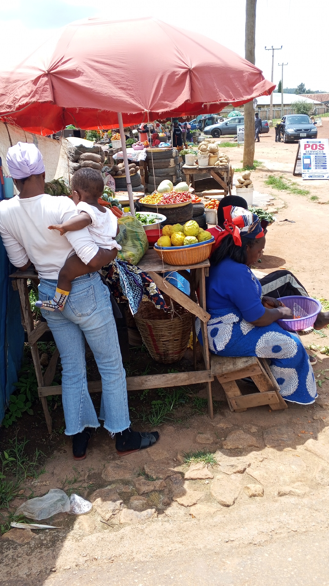 A medium shot captures a vibrant street market scene under a large, red umbrella in Jos, Nigeria. A woman, viewed from behind, is holding a young child who is resting on her shoulder. She is wearing a white t-shirt and light blue flared jeans. The child is dressed in a white garment. To the right, a woman in a blue dress with a white circular pattern and a matching headscarf is seated on a wooden stool. She is actively engaged in sorting items in a purple bowl. The market stall, constructed from wood and tires, displays an abundance of produce, including large platters of red and yellow peppers, green leafy vegetables, and yellow fruits in an orange basket. Various baskets and plastic bags are also visible. In the background, other market stalls and individuals are present, along with parked vehicles. A prominent sign advertising "POS SERVICES" stands to the right. The ground is a mixture of dirt and scattered stones.