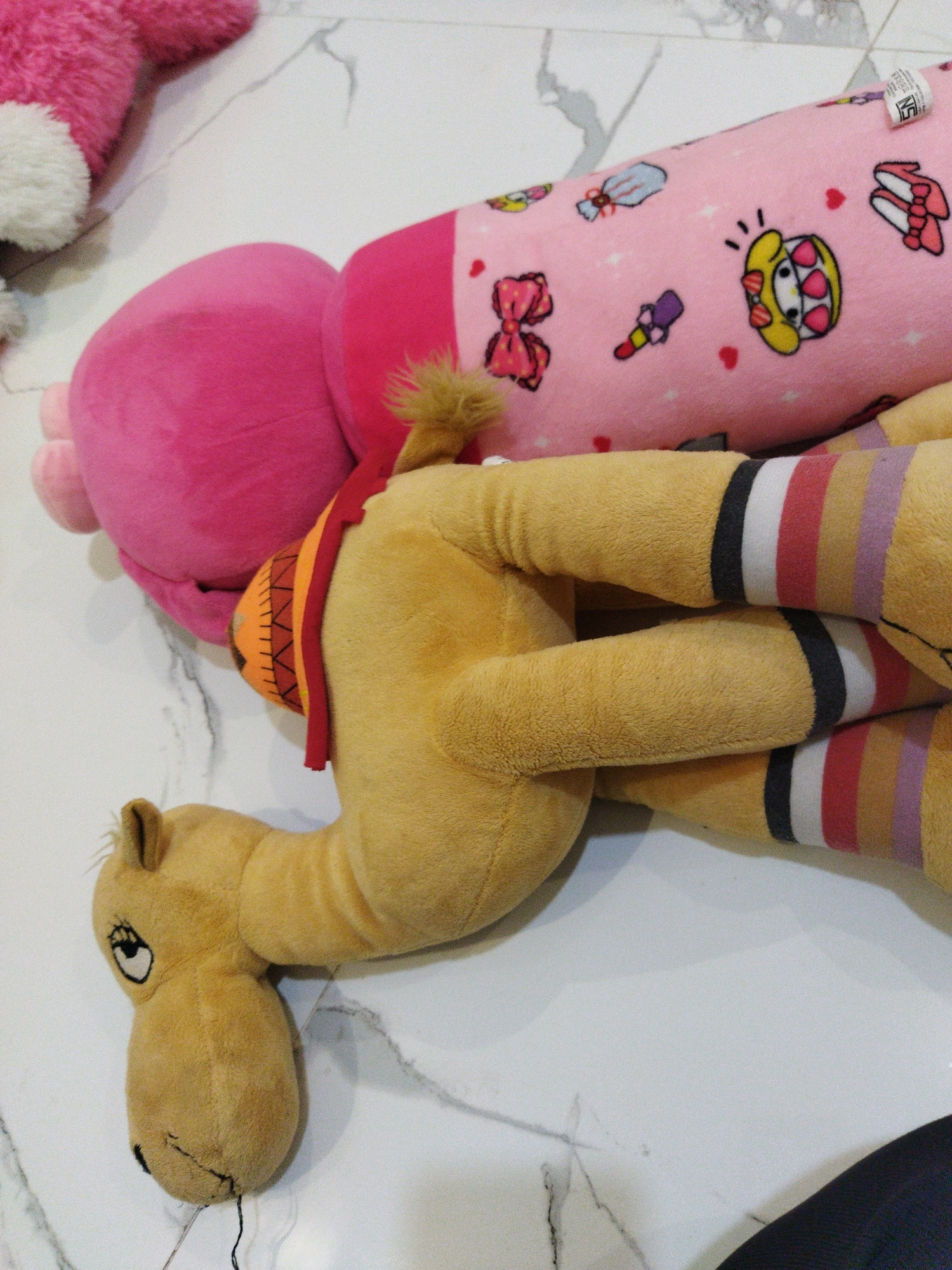 A tan stuffed camel with a red and orange saddle lies on a white marble floor. The camel is positioned horizontally, with its head facing left. Its legs are bent and crossed. Behind the camel, a large pink bolster pillow with cartoonish drawings of bows, shoes, and a character resembling Hello Kitty is visible. To the far left, a portion of a pink fuzzy object, possibly another stuffed toy, is also present. The scene appears to be indoors, likely in a residential setting. No individuals are present in the image. The location is specified as Banjar Agung, Indonesia.