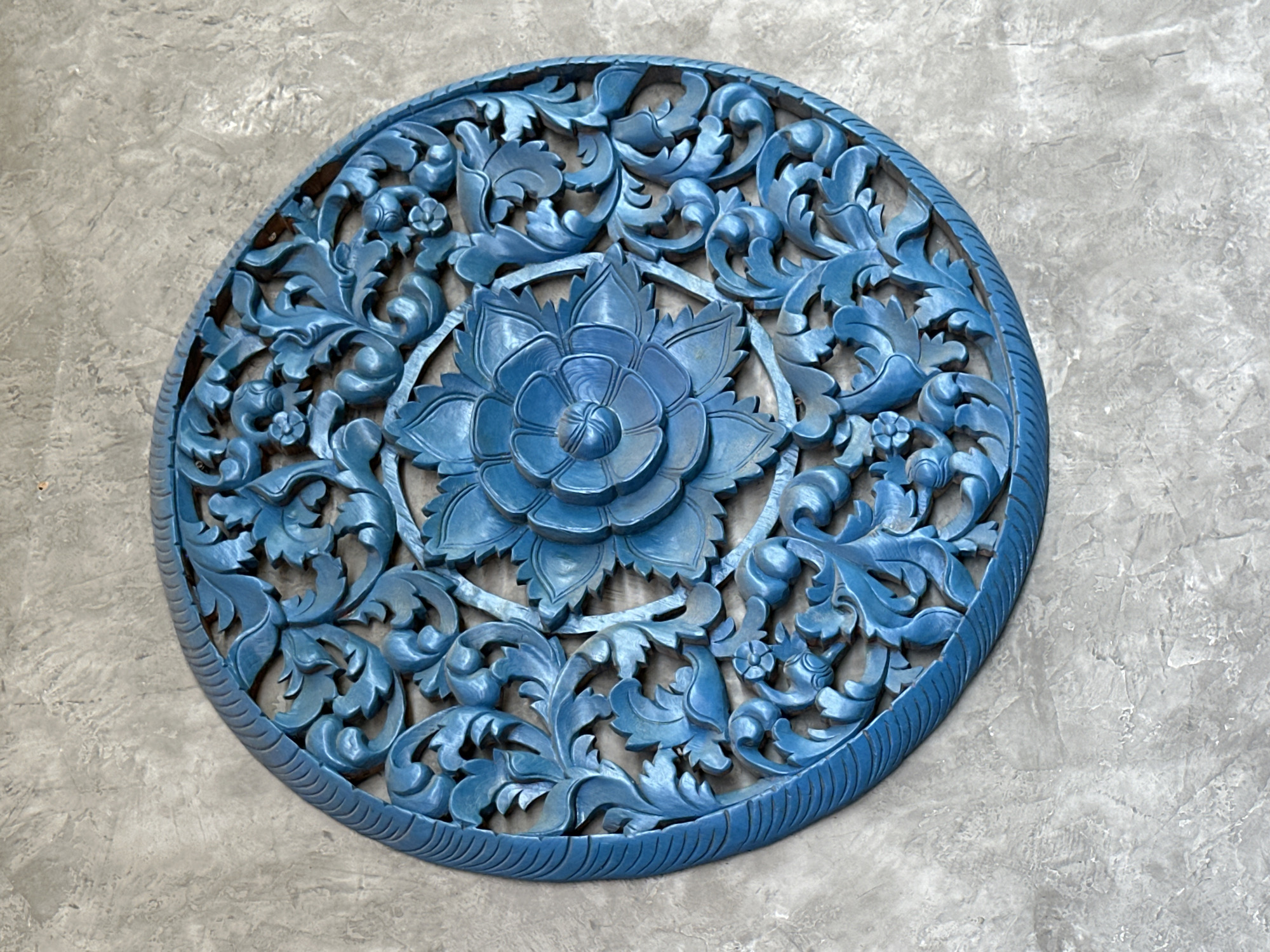 A blue, circular, intricately carved wooden art piece is mounted on a textured gray concrete wall. The carving features floral and foliate motifs, with a prominent flower at the center and radiating patterns of leaves and smaller floral elements towards the edge. The wood appears to be painted a solid, slightly matte blue. The concrete wall has a rough, uneven surface with visible variations in tone and texture, suggesting an outdoor or rustic interior setting. This art piece is located in Rawai, Thailand.