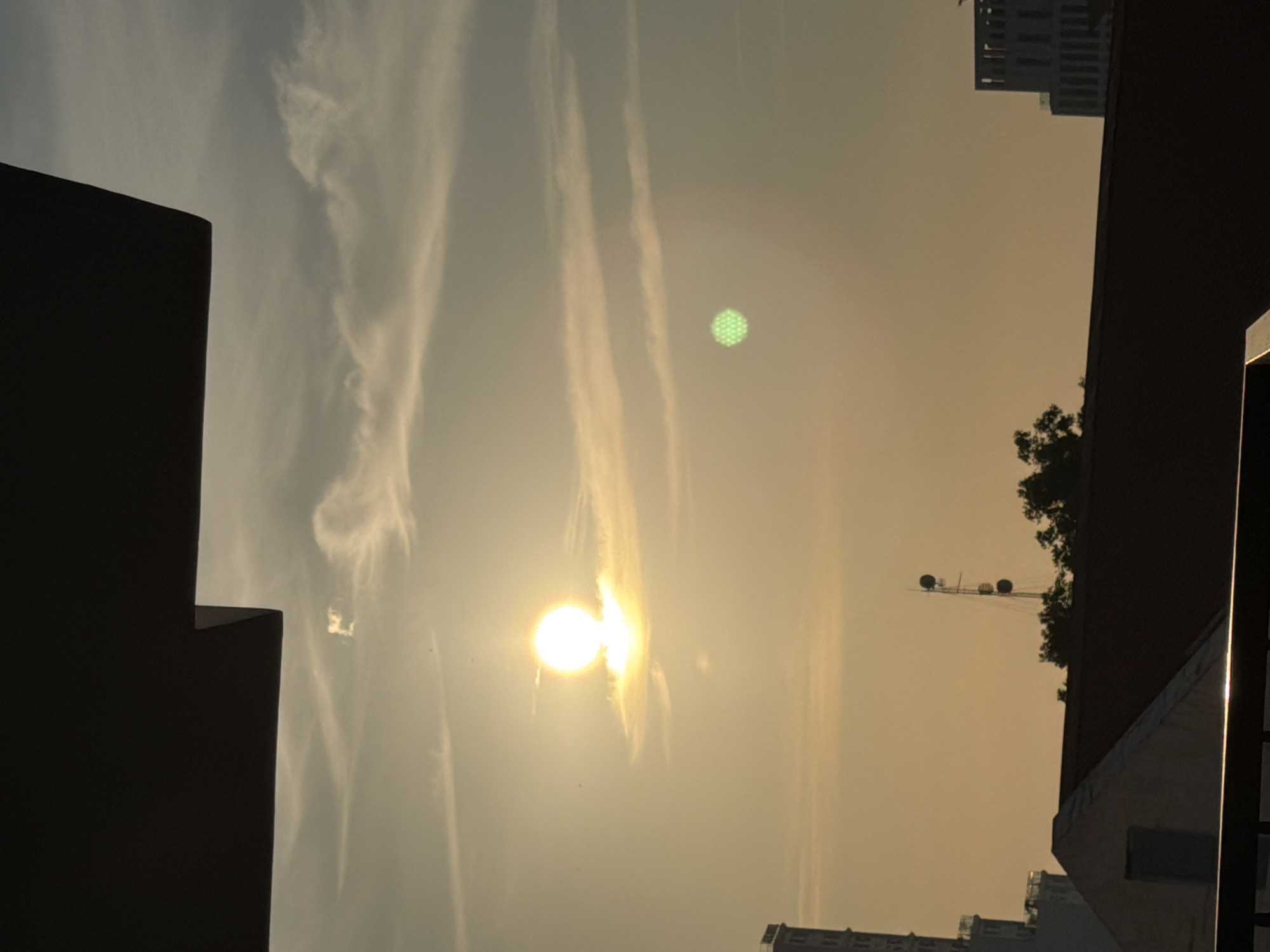 The image captures a serene sunset over the city of Nha Trang, Vietnam. The sky is a canvas of warm hues, with the sun prominently positioned in the upper left quadrant, casting a soft glow. Wispy clouds drift across the horizon, adding texture to the sky. In the foreground, the silhouettes of buildings and rooftops create a sense of depth, indicating an urban setting. A dark, angled structure, possibly an overhang or a building edge, occupies the top portion of the frame, framing the sky. A distinct circular, green object with a hexagonal pattern is visible in the middle right of the sky, possibly a lens flare or an unusual atmospheric phenomenon. The overall scene is tranquil, with no people or overt actions depicted, emphasizing the natural beauty of the sunset and the urban landscape of Nha Trang.