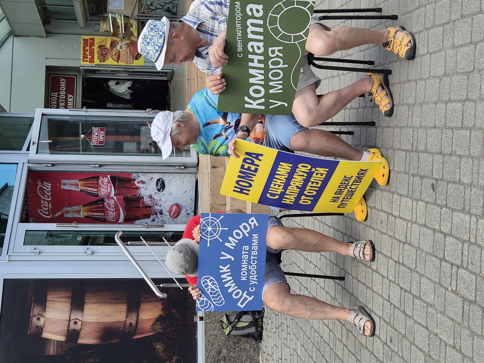 Three men sit on metal chairs on a paved walkway in Sochi, Russia. Each holds a sign advertising rooms and accommodations near the sea. The signs are printed in Russian. A store front with a Coca-Cola advertisement, a barrel design, and a sign for hot pastries is in the background.