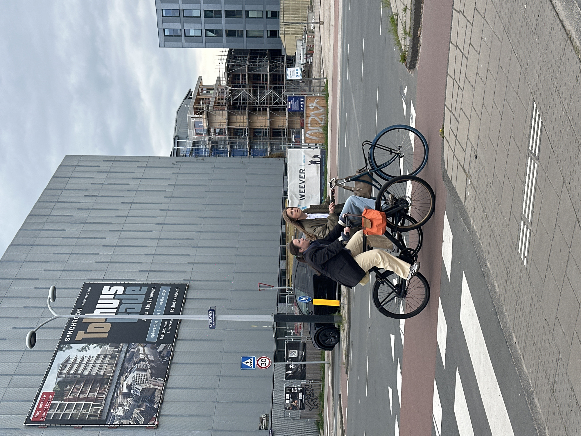 The image depicts a street scene in Amsterdam, Netherlands. Two individuals are sitting on bicycles on a crosswalk. A man, wearing a dark jacket and light-colored pants, is positioned towards the front of the image, while a woman, wearing a light jacket and jeans, sits behind him. An orange bag rests on the back of the front bicycle.
In the background, there is a large building with a billboard advertising "Tolhu's" apartments by SYNCHROON. The billboard also includes information about the apartment complex's location at Jw thuishaven, its expected delivery in mid-2024, and the website Tolhuiskade.nl. Adjacent to the building, there's a construction site with scaffolding, and a building under construction. Street signs are visible, including "ASTERWEG BUIKSLOTERHAM," a pedestrian crossing sign, and a speed limit sign indicating 30 km/h. A small sign on the construction site reads "WEEVER." A black car is parked near the sidewalk.