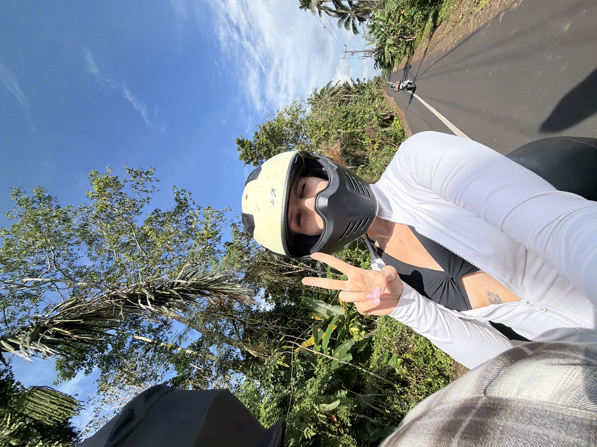 In Penebel, Indonesia, two individuals are riding on a motorcycle on a paved road surrounded by lush greenery and palm trees under a bright blue sky with scattered clouds. The person in the foreground, wearing a white zip-up jacket, a black sports bra, and a yellow helmet with a black face mask, is taking a selfie and making a peace sign with their right hand. Their fingernails are painted pink. A faint heart tattoo is visible on their chest, and a floral tattoo is on their stomach. The second person, whose head and shoulder are visible on the left, is wearing a black helmet and a plaid jacket. In the distance, another person is riding a motorcycle. Electrical wires run along the side of the road.
