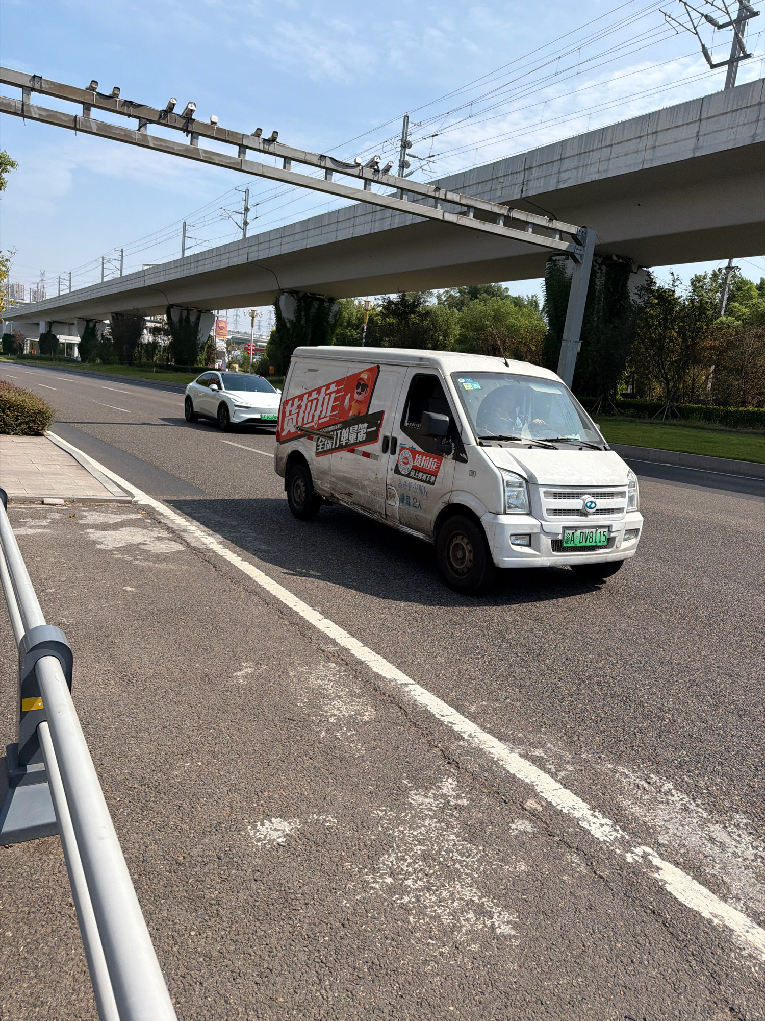 This is a low-angle, eye-level, outdoor photograph of a white cargo van with Chinese text on its side, driving on a paved road. The van has a license plate that reads "A DV8115". A white electric car with a black roof is visible in the background, also driving on the road. An elevated railway structure with overhead power lines runs across the upper portion of the image. The sky is clear and blue with some clouds. The environment appears to be a road in a city with trees and greenery on the sides. This scene is located in Jiangjin District, Chongqing, China, near the Jiangjin Yangtze River Bridge.