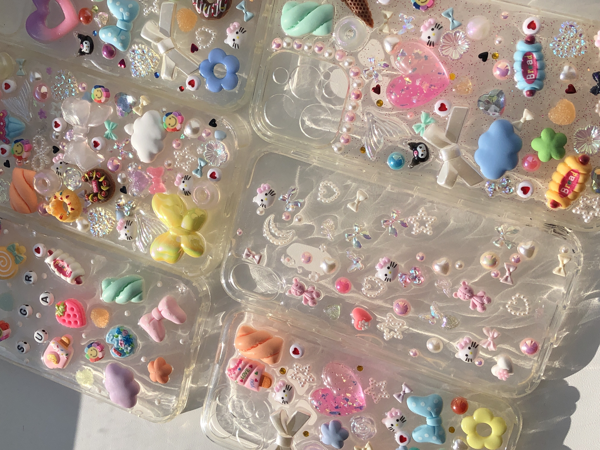 The image shows a collection of six decorated phone cases arranged on a white surface. The cases are transparent and embellished with various miniature ornaments, including faux sweets, floral motifs, bows, hearts, and character figurines. The light source is strong and creates shadows, indicating an outdoor setting. No people or actions are visible in the image. The location is not discernible from the provided image.