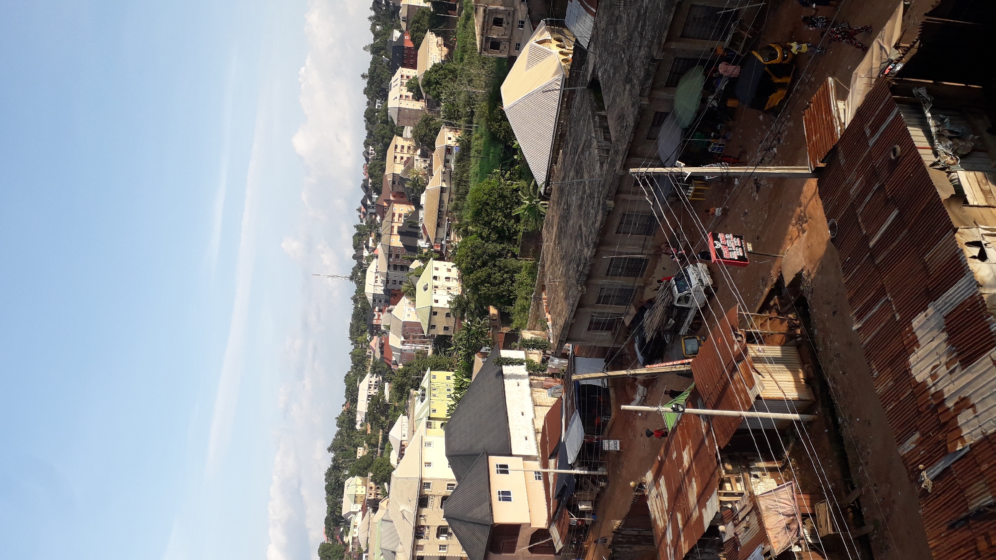 This is a high-angle, vertical, full shot of a bustling street scene in Nkpor, Nigeria. The sky is a pale blue with scattered white clouds. The foreground is dominated by rustic, corrugated iron roofs, some showing signs of rust and decay. A dirt road winds through the scene, lined with various buildings and structures.
In the mid-ground, the road is busy with activity. A white pickup truck is parked on the side, and a yellow and black tricycle, a common mode of transport in Nigeria, is in motion. Several people are visible on the street and sidewalks, engaged in daily activities. Some appear to be vendors or shoppers, with stalls and makeshift shops set up along the road. A prominent red and white sign with text is mounted on a pole.
The background reveals a densely populated urban landscape with a mix of residential and commercial buildings. The architecture varies, with some multi-story structures painted in light colors and others appearing more basic. Lush green trees and vegetation are interspersed among the buildings, indicating a tropical environment. A cell tower is visible in the distance on a hilltop. The overall impression is one of a vibrant and active community.