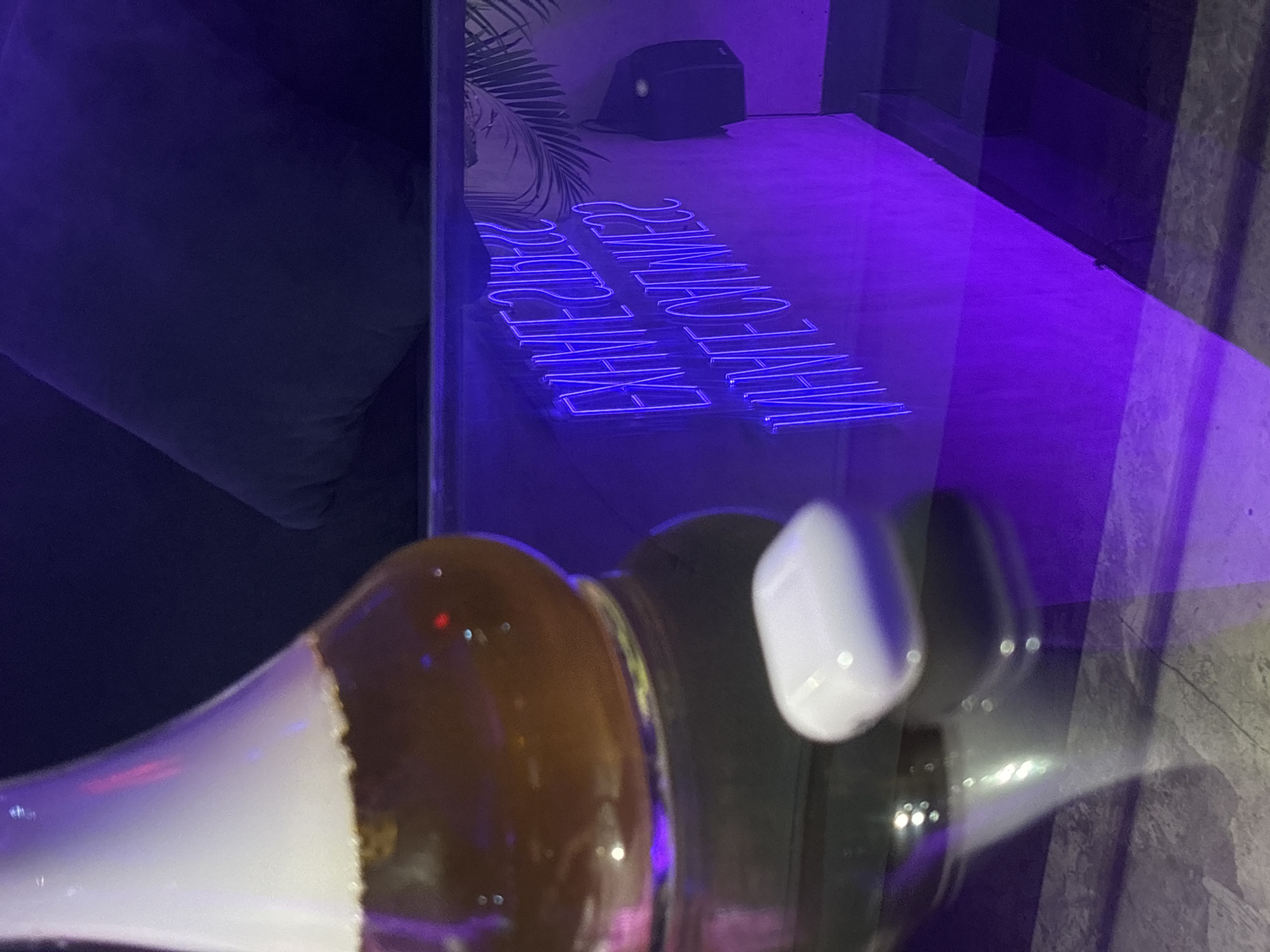 The photograph presents a scene captured in what appears to be a dimly lit indoor environment. Reflected in a glass surface is a blue neon sign that reads "Exhale Vibes" and "Inhale Greatness" but is mirrored. A dark object, possibly a speaker, is placed on a surface near the neon sign and a large plant. In the foreground, out of focus, are objects that appear to be a glass bottle and white wireless earbuds, possibly Apple AirPods. The location, Kuta Selatan, Indonesia, is not verifiable from the visual information in the image.