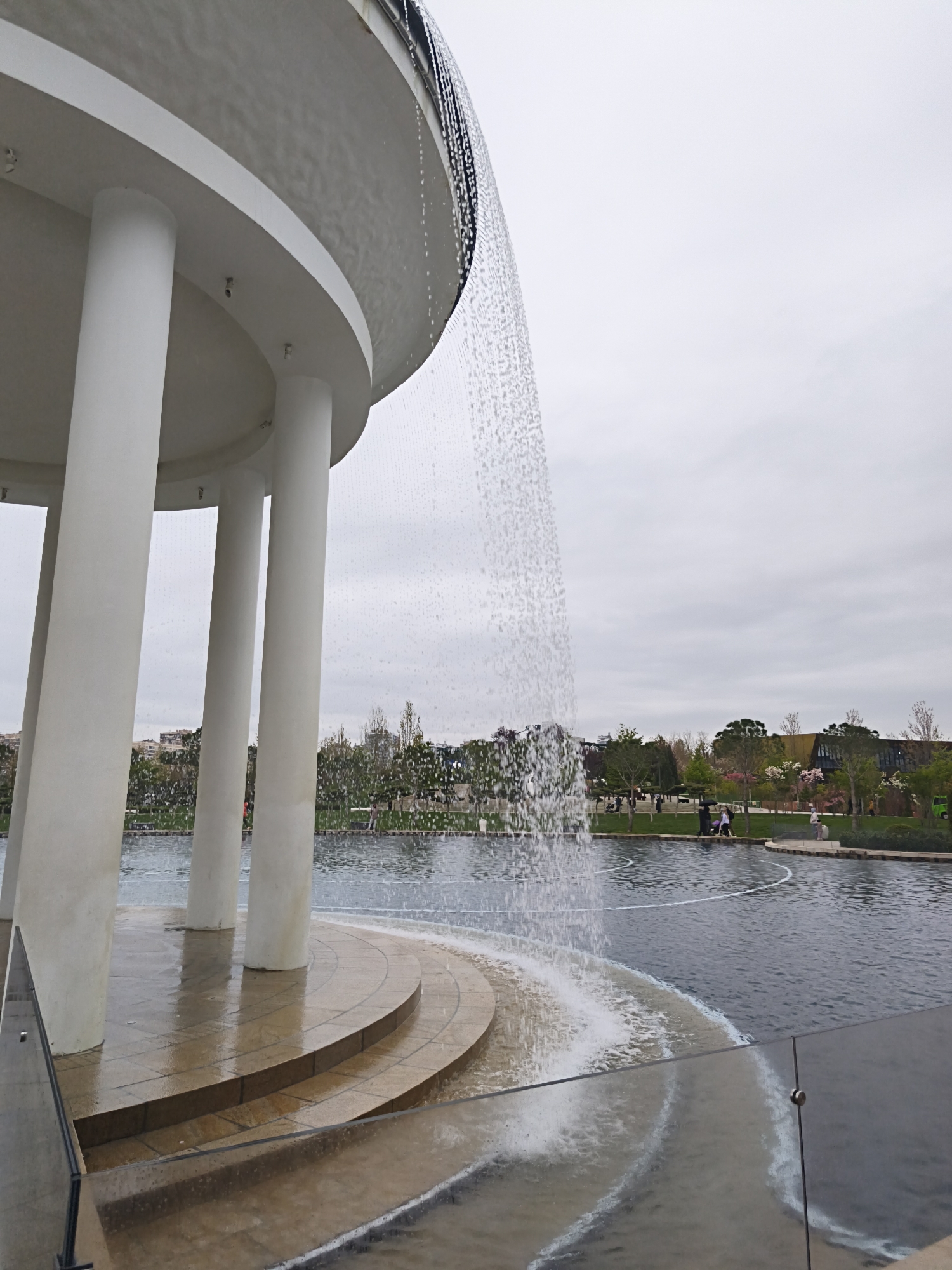 The image depicts a circular, colonnaded structure with water cascading from its upper edge into a pool. The structure has a raised, tiered base of beige tiles. The pool is dark and reflects the sky. In the background, trees and shrubs line the edge of the pool. Several people are visible along the pool's edge, appearing to be strolling or resting. The sky is overcast. The location is the Galitsky Park in Krasnodar, Russia.