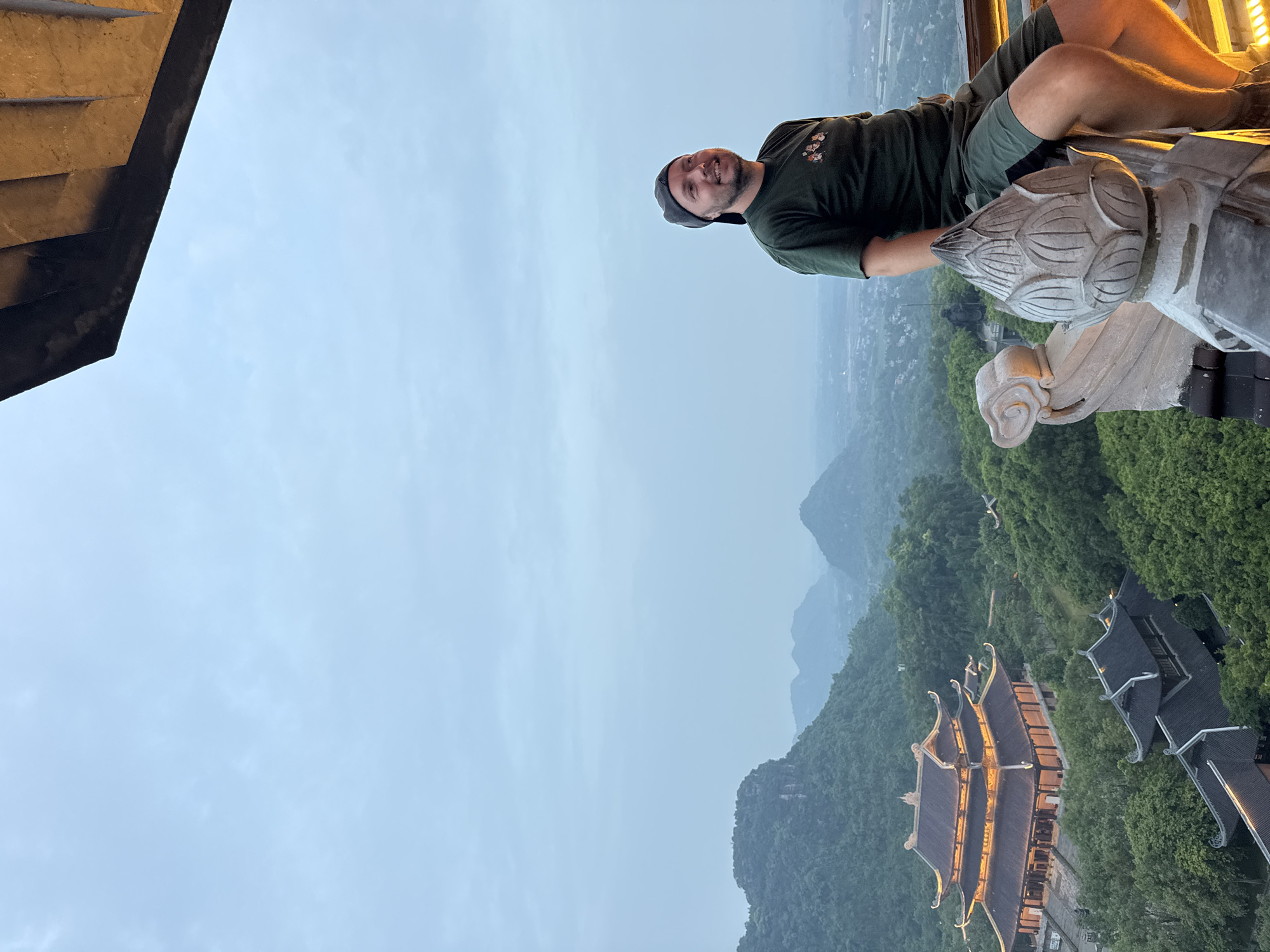 A man wearing a dark green t-shirt and shorts, and a dark baseball cap is sitting on a ledge overlooking a valley. The man is looking to his left and smiling. In the background, there is a large temple complex with illuminated roofs nestled among lush green trees. Mountains are visible in the distance under a hazy sky. The temple complex appears to be located in the city of Tây Hoa Lư Ward, Vietnam, known for its historical and cultural significance, including ancient temples and pagodas. The architecture suggests a traditional East Asian style, with ornate roofs and pagodas. The overall scene depicts a scenic view of a temple in a mountainous region, possibly at dusk or dawn given the lighting conditions.