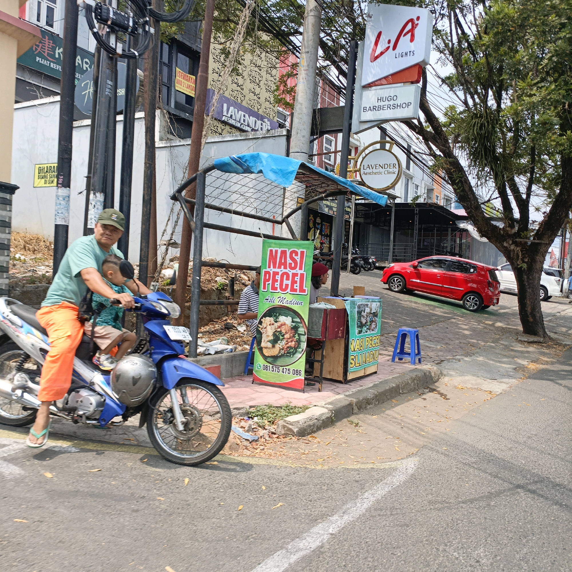 The image depicts a street scene in Salatiga, Indonesia, likely on a sunny day. In the foreground, a man wearing an olive green t-shirt and bright orange pants is riding a blue and silver motorcycle. He is wearing a green baseball cap and has a child seated in front of him, also wearing a t-shirt and shorts. The child appears to have a dark object attached to their head. A silver helmet rests on the front of the motorcycle.
To the right of the motorcycle, a street vendor stall is visible. A tall green sign with red lettering reads "NASI PECEL Asli Madiun" and features a circular image of the dish. Below this, a phone number "081 575 475 086" is displayed. Next to the sign is a wooden cart with glass panels displaying more food items. A small blue plastic stool is placed near the cart.
In the background, buildings line the street. One building has a prominent red and white sign that says "L.A. LIGHTS" and below that "HUGO BARBERSHOP". Another sign reads "LAVENDER Aesthetic Clinic". There are several utility poles with a dense network of wires running between them. A red car is parked on the street to the right, and a white car is further down the road. A large tree with dense foliage is situated on the right side of the image, casting shadows on the street. The ground is paved with asphalt and some stone or concrete curbing is visible. Debris, including leaves and what appears to be blue plastic, is scattered along the side of the road. The overall impression is a bustling urban environment with commercial activity.