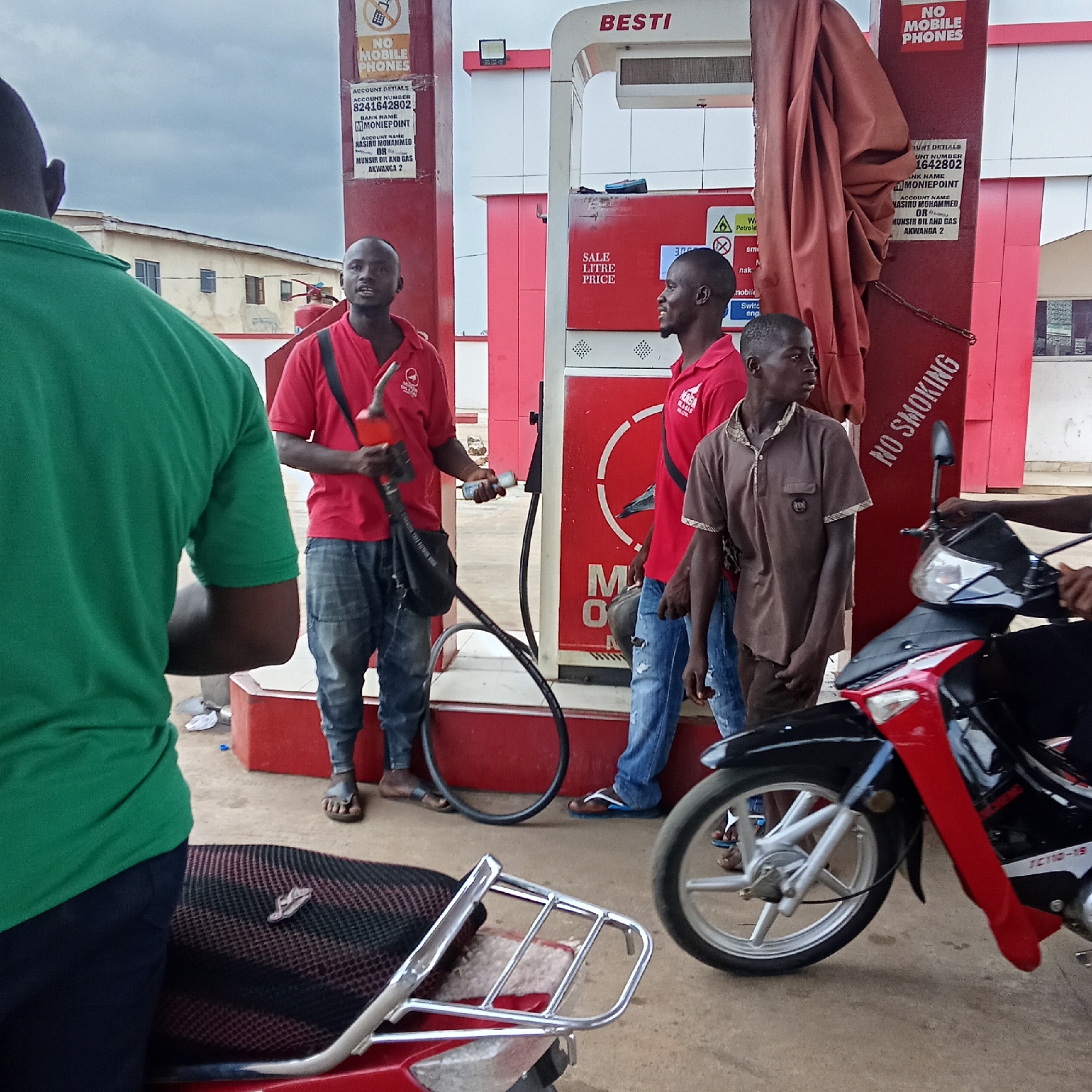 This medium-shot image captures a scene at a gas station in Akwanga, Nigeria, under an overcast sky. The central focus is a red and white fuel pump, branded "BESTI," with several individuals engaged in activity around it.
In the foreground, a man wearing a green t-shirt stands with his back to the camera, partially obscuring the seat and chrome rack of a red motorcycle. To his right, a fuel attendant in a red polo shirt, featuring a "MUNSA OIL & GAS" logo, holds a fuel nozzle and hose, appearing to be in the process of refueling. He looks towards the viewer. Another fuel attendant, also in a red polo shirt, stands to the right of the pump, looking towards the right. A young man in a brown polo shirt stands between this attendant and the pump. On the far right, the front portion of a red and black motorcycle, marked with "JC110-19," is visible, suggesting it is next in line or being refueled.
The fuel pump's display shows "SALE LITRE PRICE" and a partial numerical value, "300." Multiple signs are affixed to the pump structure. These include "NO MOBILE PHONES" and "NO SMOKING" warnings for safety. Another sign provides "ACCOUNT DETIALS" for payment, listing "ACCOUNT NUMBER 8241642802," "BANK NAME MONIEPOINT," and "ACCOUNT NAME NASIRU MOHAMMED OR MUNSIR OIL AND GAS AKWANGA 2." A red fire extinguisher is visible in the background, near a building. Most individuals are wearing sandals or flip-flops. The scene depicts a routine transaction at a local gas station.