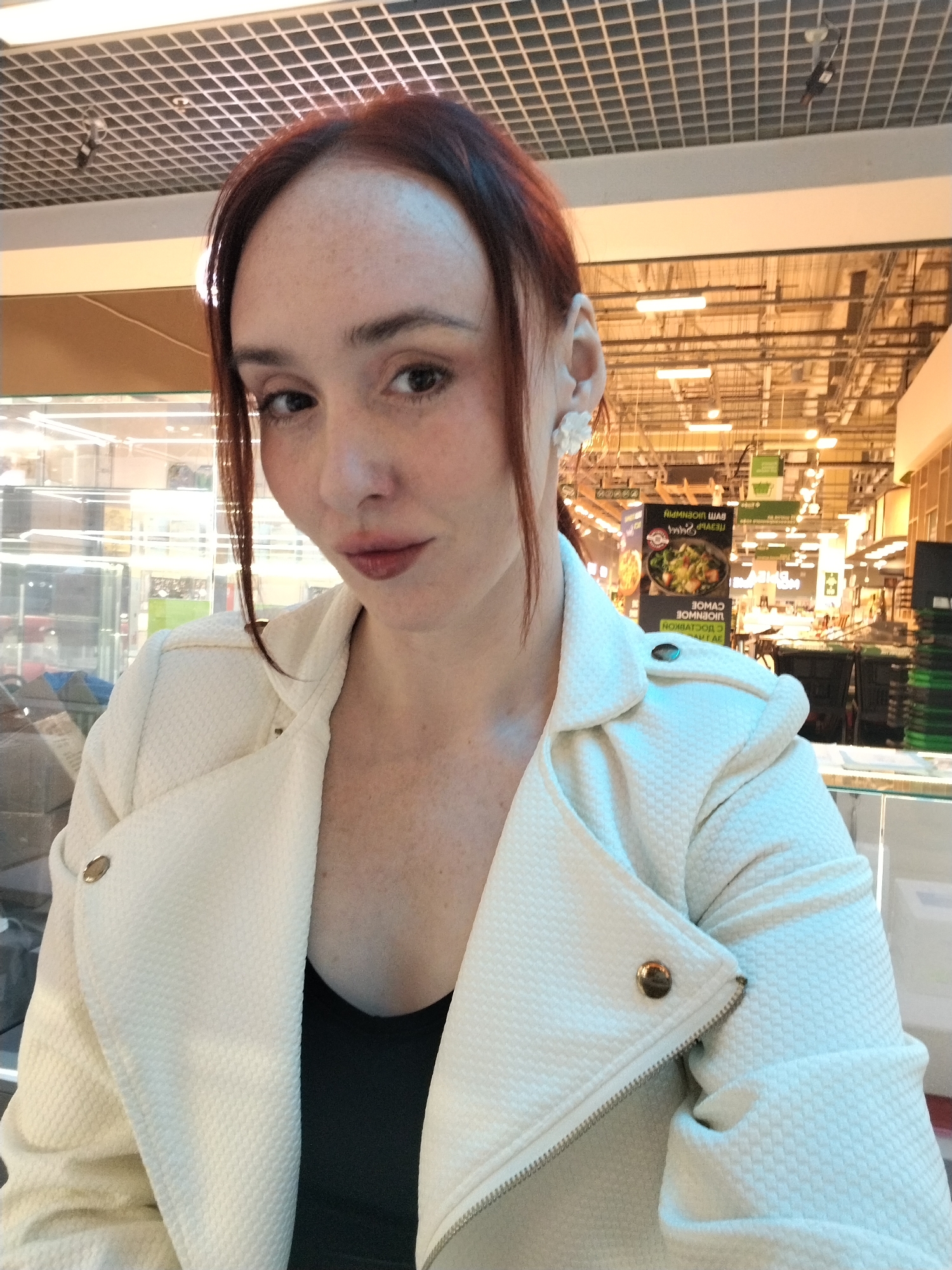 A woman is posing for a selfie inside what appears to be a supermarket or shopping mall in Izhevsk, Russia. She has reddish-brown hair, fair skin with freckles, and is wearing a white textured jacket over a black top. She is facing the camera, and the background shows supermarket aisles, overhead lighting structures, and informational signs with Russian text.