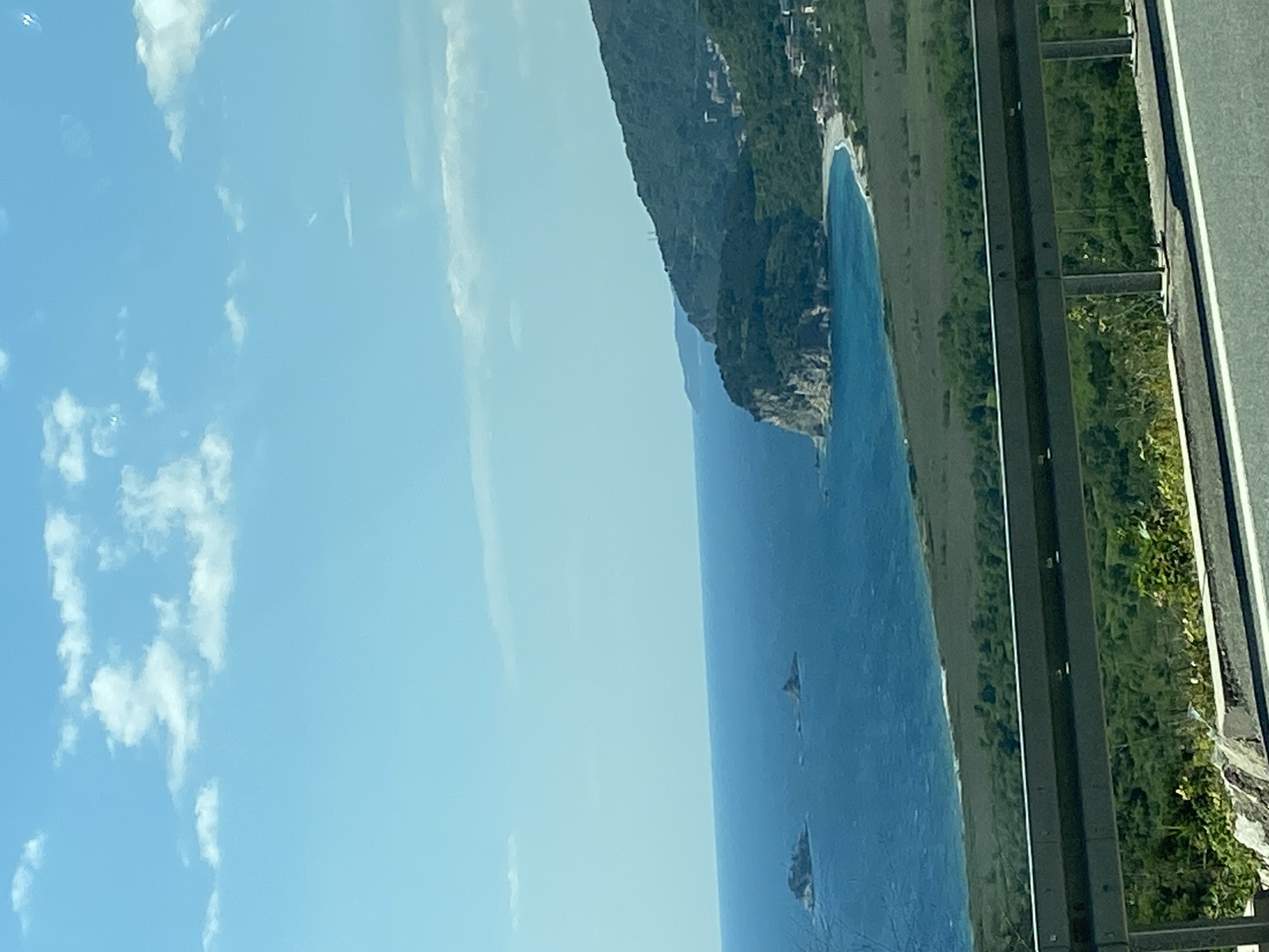 The image, captured in a portrait orientation and rotated 90 degrees clockwise from its natural landscape view, depicts a stunning coastal panorama. The scene is viewed from an elevated position, likely a roadside, with a grey asphalt road and a metallic guardrail visible on the right side of the frame (when the image is correctly oriented).
The dominant features are the deep blue sea, a rugged, forested coastline, and a clear sky with scattered clouds. The tranquil expanse of the sea occupies the lower half of the correctly oriented image, dotted with a few dark, rocky islets emerging from the water. A prominent, verdant mountain or cliff face, densely covered in green vegetation, forms the coastline, extending from the middle ground towards the horizon. At the base of these cliffs, a crescent-shaped, light-colored beach or cove is visible, where the calm water gently meets the shore. Faint details of structures or buildings can be seen nestled within the greenery further up the slopes, suggesting a coastal settlement.
The sky, occupying the upper portion, is a bright, clear blue with soft, white cumulus clouds. The lighting indicates a bright daytime setting, possibly late morning or early afternoon, with warm sunlight illuminating the landscape. The overall atmosphere is one of serene natural beauty. No people or visible text are present in the image. The rugged topography, clear blue waters, and green vegetation are characteristic of coastal regions, consistent with the specified location of Kanjoši, Montenegro.