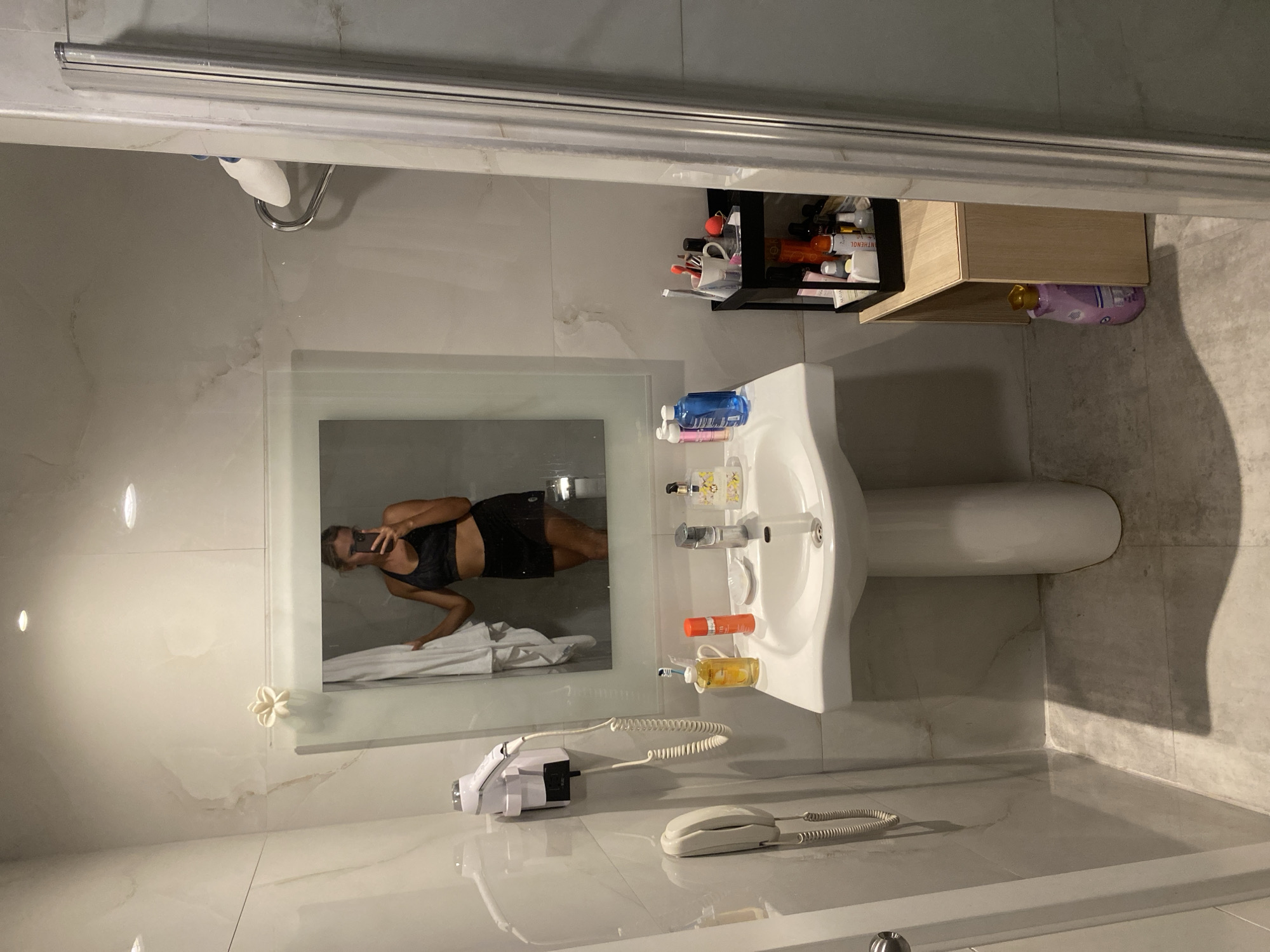 A young Caucasian woman with blonde hair is taking a mirror selfie in a modern bathroom. She is wearing a black sports bra and black shorts. The bathroom features a white pedestal sink, a large mirror, a wall-mounted hairdryer, and a shower enclosure. Various toiletries and cosmetics are visible on the sink counter and a small shelf unit to the right of the sink. The walls are tiled with a marble-like pattern, and the floor is tiled with large grey tiles. The scene is indoors, in what appears to be a hotel room or a residential bathroom. The location of Aksu, Turkey is not discernible from the image.