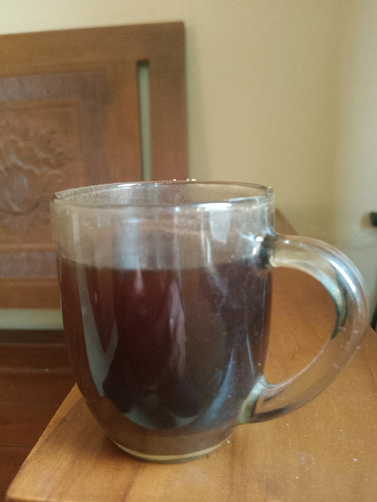 A clear glass mug filled with a dark liquid, likely coffee or tea, sits on a wooden surface. The mug has a clear glass handle. In the background, a piece of furniture, possibly a wooden cabinet or chair, is visible. The overall scene appears to be indoors, with a plain wall in the background. The lighting is soft, suggesting natural light from a window or ambient room lighting. The image is taken from a slightly elevated angle, looking down at the mug. There is no indication of the specific location within Sleman, Indonesia, beyond the user's prompt.