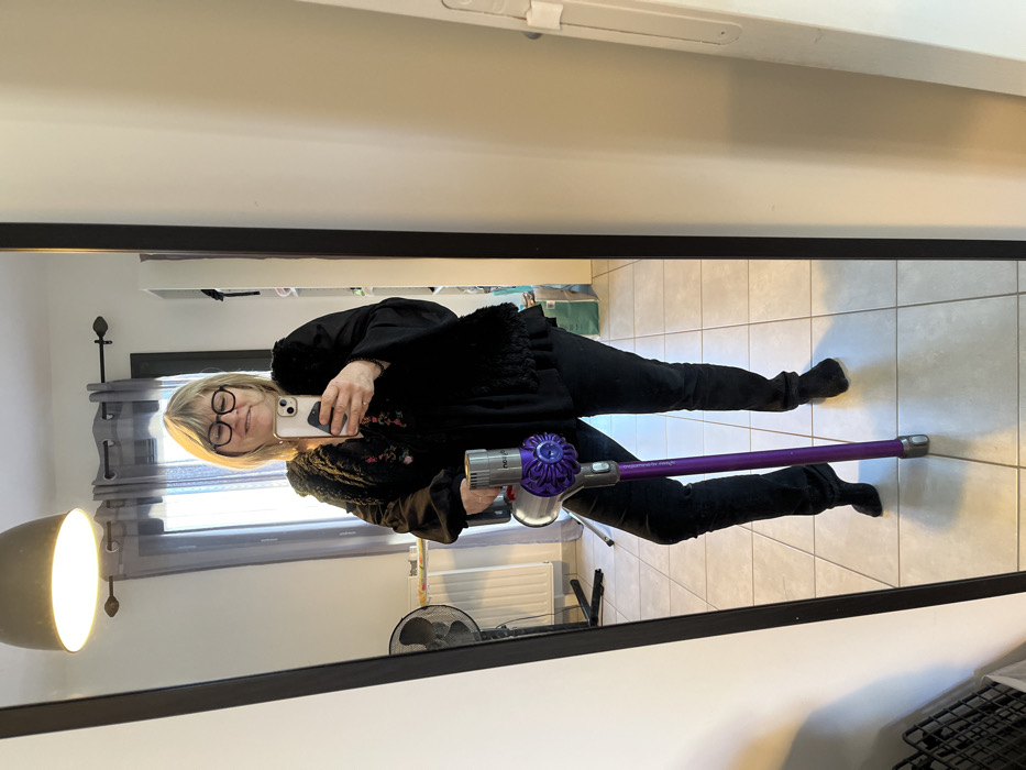 A blonde, middle-aged woman is depicted in a full-length mirror selfie taken in a room located in Chambœuf, France. She wears dark-framed glasses, a black long-sleeved shirt with a black faux fur vest, black trousers, and black ankle boots. Her right hand holds a light-colored smartphone, capturing the image, while her left hand supports a silver and purple Dyson cordless vacuum cleaner leaning against her left leg. She presents a smiling expression.
The room's interior, visible in the reflection, features a light-tiled floor and light-colored walls. A large window with dark grey curtains on a dark rod is centered in the background, with a white radiator below it and a black standing fan to the left. An illuminated bell-shaped ceiling light fixture is visible overhead. A white shelving unit, a light blue bag on the floor, and a dark wire basket are partially visible elements within the reflected space.