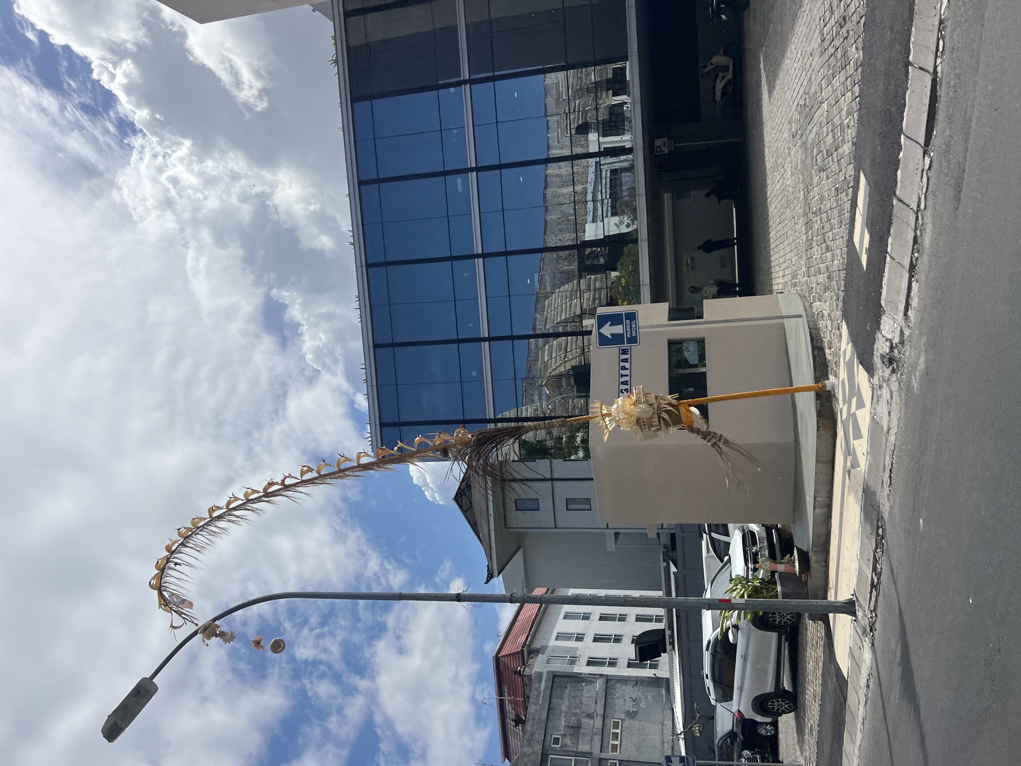 In Kuta, Indonesia, on a paved street, a street lamp with a decorative, dried palm frond arching over it stands near a modern building with large, reflective blue windows. A beige, low-rise structure with the word "SATPAM" (security) on it is situated in front of the modern building. A blue sign with an upward arrow and the text "PARKIR MOBIL" (car park) is mounted on this structure. Several cars, including a white SUV and a black sedan, are parked on a cobblestone area adjacent to the structures. The sky is cloudy with patches of blue. The scene is outdoors during the daytime.