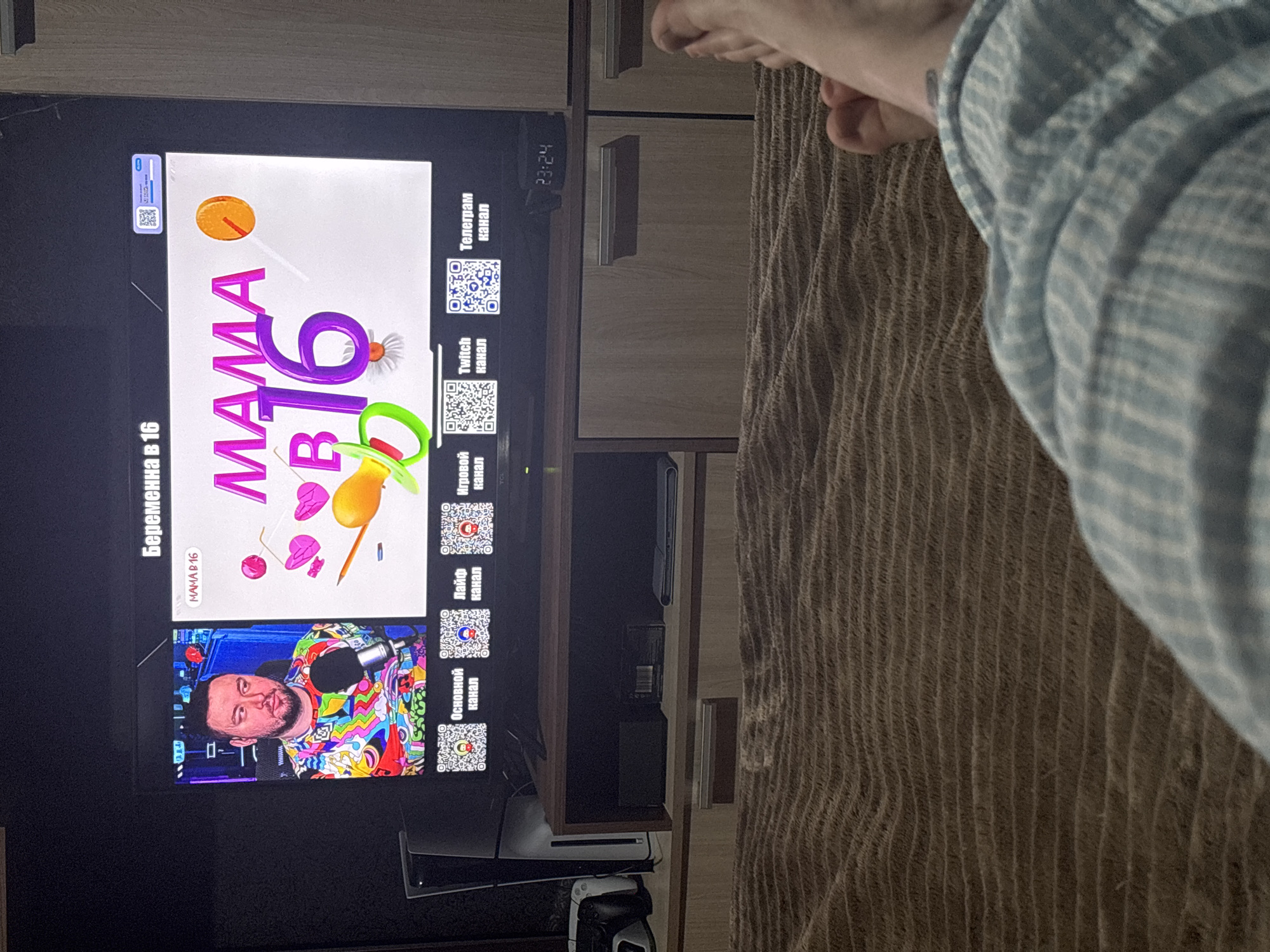 A television displays a show titled "MAMA B 16" in Russian, also labeled "Pregnant at 16." The screen features graphics and a live-stream of a man with a microphone, dressed in a colorful shirt. QR codes are placed on the right side of the screen, presumably for channel links. The TV is set in a room with wooden furniture. The time displayed on the screen is 23:24. A person's foot and leg, covered by blue and white checkered pajamas, are visible in the upper right corner of the image. A console and other devices are on a shelf under the TV. The location is Chornomorskoe, Ukraine.