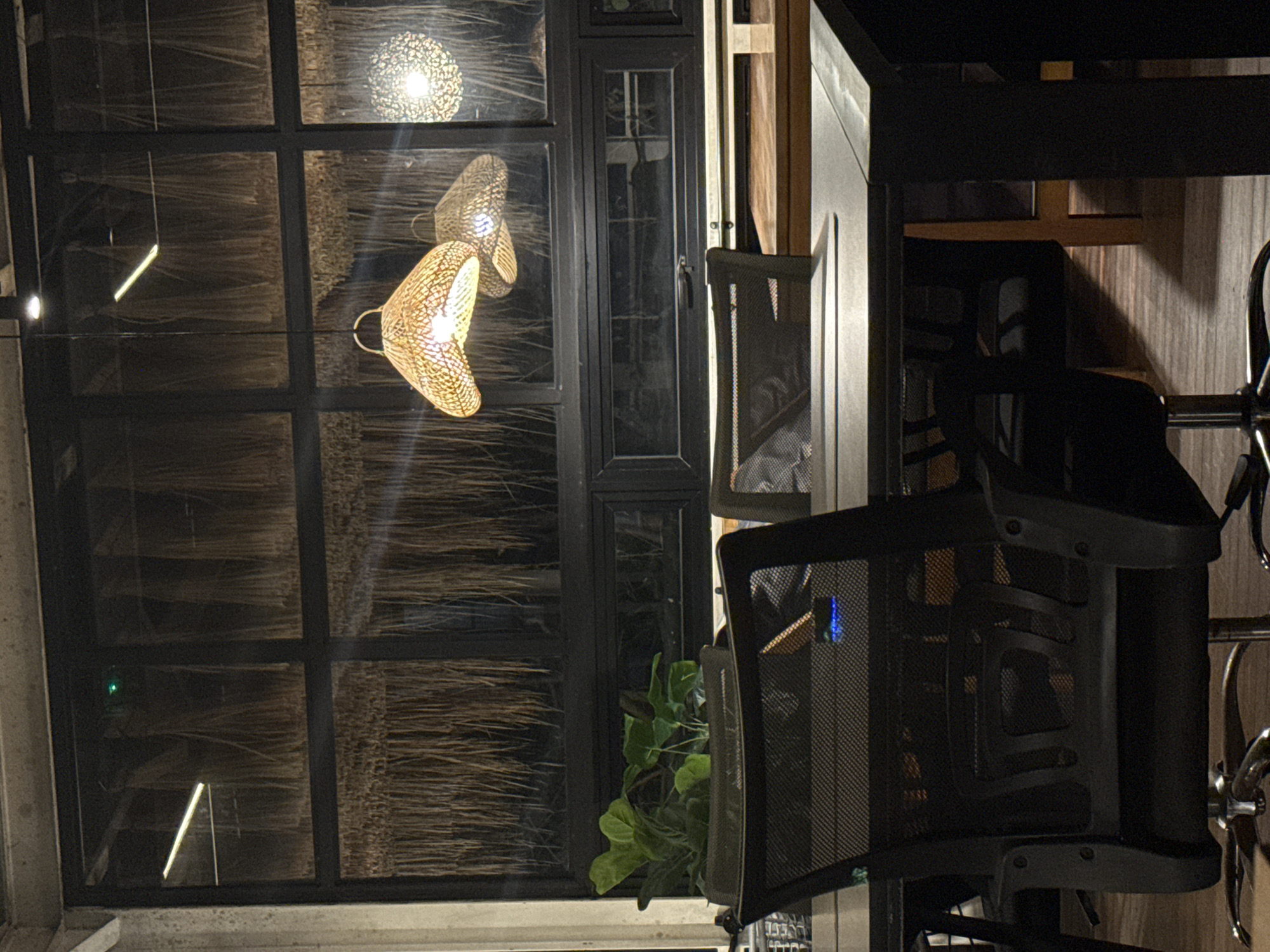 This image depicts an indoor scene with a prominent window that looks out onto a thatched roof or wall. Through the window, two woven pendant lights with a warm, yellow glow are visible, hanging from the ceiling. One light is spherical and patterned, while the other has a more organic, teardrop shape. The window itself is divided into multiple panes by dark frames.
In the foreground, a dark mesh office chair with a metallic base and armrests is partially visible. Another similar chair is seen to the right, with its chrome accents catching the light. A green potted plant with large, broad leaves is positioned near the window, adding a touch of life to the scene.
The lighting suggests it is nighttime or dusk, with artificial lights providing illumination indoors. The overall atmosphere is cozy and potentially part of a restaurant, cafe, or lounge area. The view through the window, with the thatched material, hints at a tropical or beachside location, consistent with Kuta Selatan, Indonesia. No people are visible in the frame. There is no visible text.