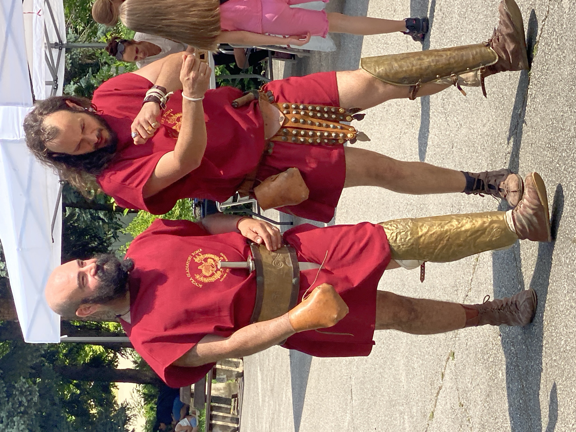 The image displays two male individuals, dressed in historical Roman-style attire, standing on a paved outdoor surface during daylight. The setting is identified as Pleven, Bulgaria.
The man on the left has a bald head and a prominent dark beard, smiling and looking towards his right. He wears a short-sleeved, knee-length red tunic. The tunic's chest features a circular gold emblem with red accents, bearing the text "SCUOLA GLADIATORI ROMA" and "GRUPPO DEI CITTADINI ROMANI". A wide brown leather belt with a sword sheath and hilt is secured around his waist. His right hand is encased in a light brown leather protective glove or covering. A golden greave (shin guard) is visible on his left leg, and he wears brown lace-up shoes.
The man on the right has long, dark, wavy hair and a beard. He is observed looking down at his left forearm, which is adorned with multiple dark and light-colored bracelets; a silver ring is visible on his left ring finger. He wears a short-sleeved, knee-length red tunic. From a belt, a small brown leather pouch hangs on his right hip, and a larger, studded brown leather fringed element hangs on his left hip. A golden greave is fitted on his right leg, and he wears brown lace-up shoes similar to the first man.
In the background, a white canopy tent is erected. Lush green trees and bushes are visible beyond the tent. Other individuals are present: a woman with sunglasses atop her head is positioned behind the second man, and a young girl with long light hair, dressed in a pink top and shorts, stands to the right of the frame. Further back, people are seated on wooden benches. The ground is a light-toned paved surface exhibiting cracks and seams. The scene is illuminated by strong sunlight, casting visible shadows.