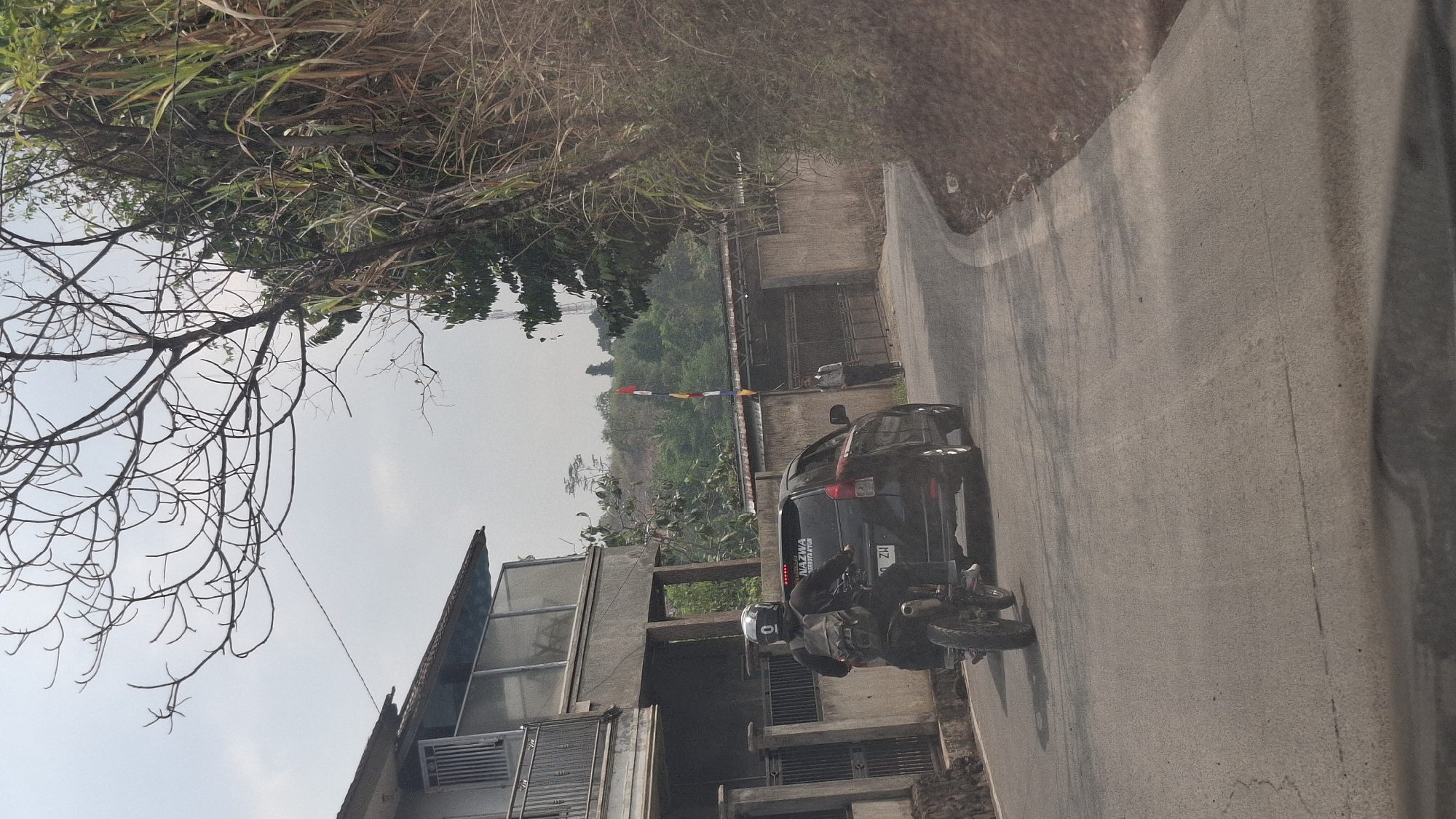 In the city of Sirnagalih, Indonesia, a motorcycle carrying a person with a backpack is traveling on a paved road. To the right of the motorcycle, a black car is also moving forward. A person is standing near a gated entrance on the right side of the road, next to a concrete wall. A building with balconies and windows is visible on the left. The road curves to the right, with a hillside covered in vegetation on the right side. The sky is overcast, and there are trees and distant hills in the background. A flagpole with colorful flags is visible in the middle distance.