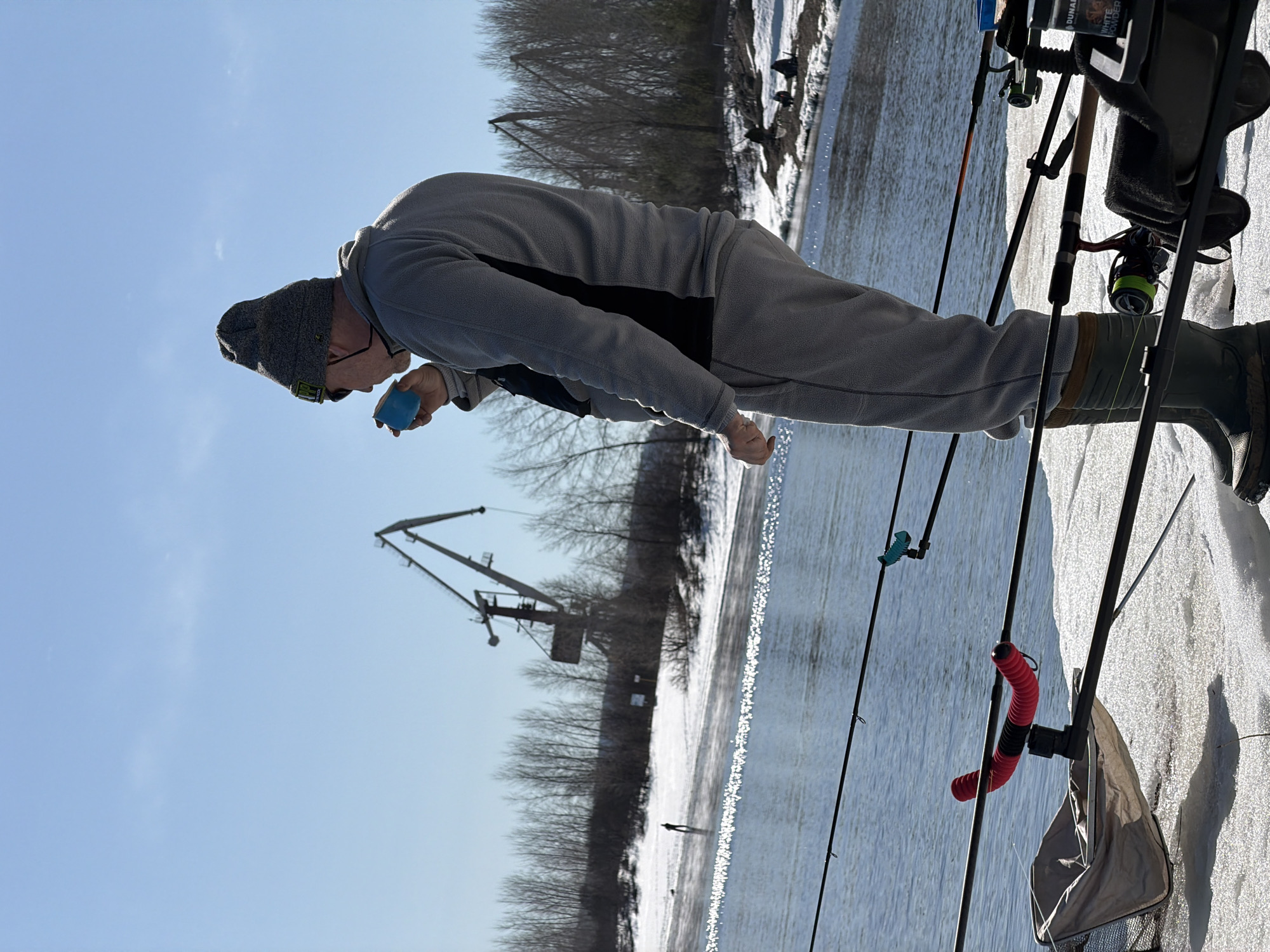 The image shows a man standing on a snow-covered bank with several fishing rods set up, likely ice fishing. He is wearing a grey fleece suit, a dark beanie, glasses, and rubber boots, and is tilting a small blue cup towards his mouth, possibly drinking. The background includes a body of water with visible ripples, bare trees, and an industrial crane on the opposite bank. The location is inferred to be near the city of Chaikovsky, Russia, based on the presence of the crane and general wintery landscape.