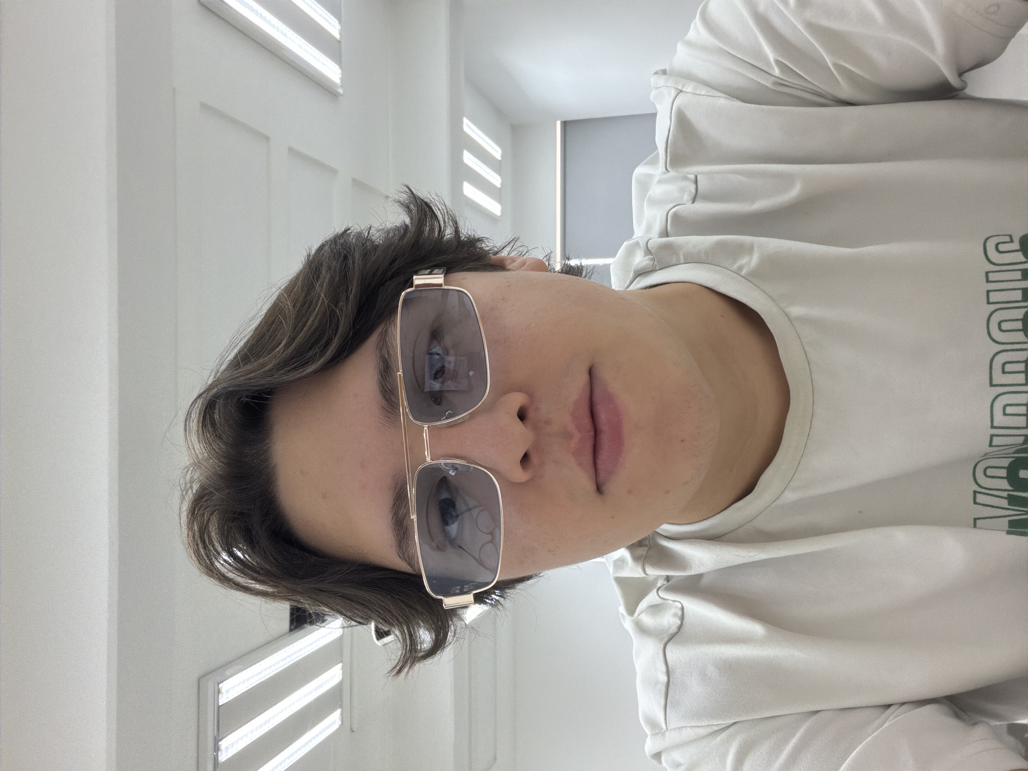 The image presents a frontal, chest-up view of a young individual, likely male, with fair skin, a neutral expression, and dark brown, wavy hair. The subject wears gold-framed, rectangular sunglasses with blue-tinted lenses that reflect several overhead fluorescent light strips. A light-colored (possibly off-white or light beige) crew-neck t-shirt with a partial green word, "VONDRON," visible on the lower front is worn. The background consists of a plain white ceiling with multiple rectangular fluorescent light fixtures, including one set of three parallel strips and another set of four. A light gray wall or partition is visible in the right background. The scene takes place indoors, likely within an institutional or commercial building, in San Andrés Cholula, Mexico.