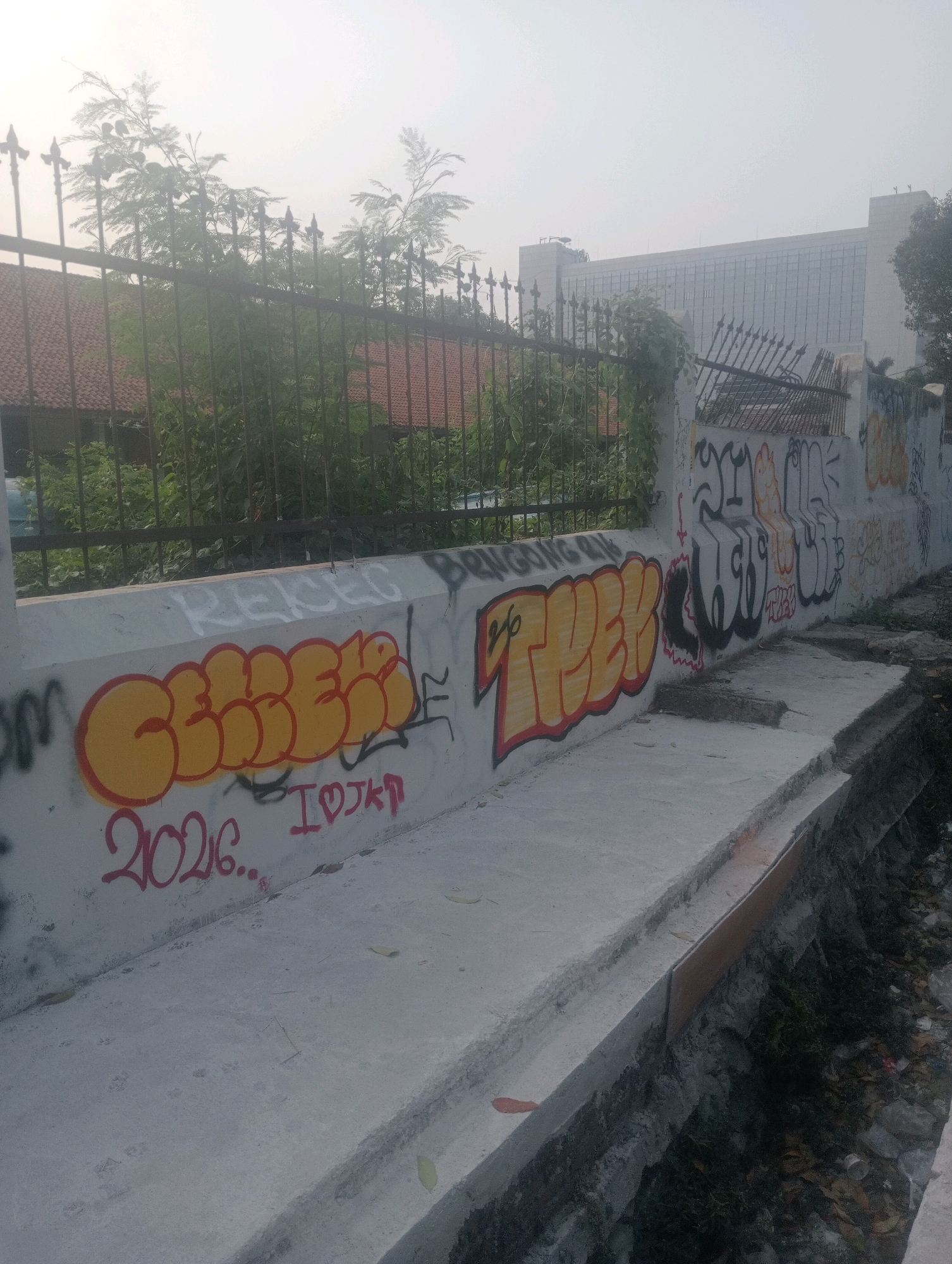 The image depicts a concrete wall with various graffiti tags. On the left, a large yellow bubble-style graffiti tag with a red outline reads "GBBBB". Below it, in pink, is "2026...". To the right of "GBBBB", another yellow and red graffiti tag reads "TPBY". Further to the right, more graffiti in black, white, and orange can be seen, including what appears to be stylized letters. A black metal fence runs along the top of the wall, with greenery and a building visible behind it. The wall itself appears to be part of a drainage system, with a concrete channel running alongside it. The sky is hazy, suggesting either a foggy or smoggy atmosphere. The graffiti suggests an urban environment, possibly a street or an underpass, in East Jakarta, Indonesia, based on the context provided.