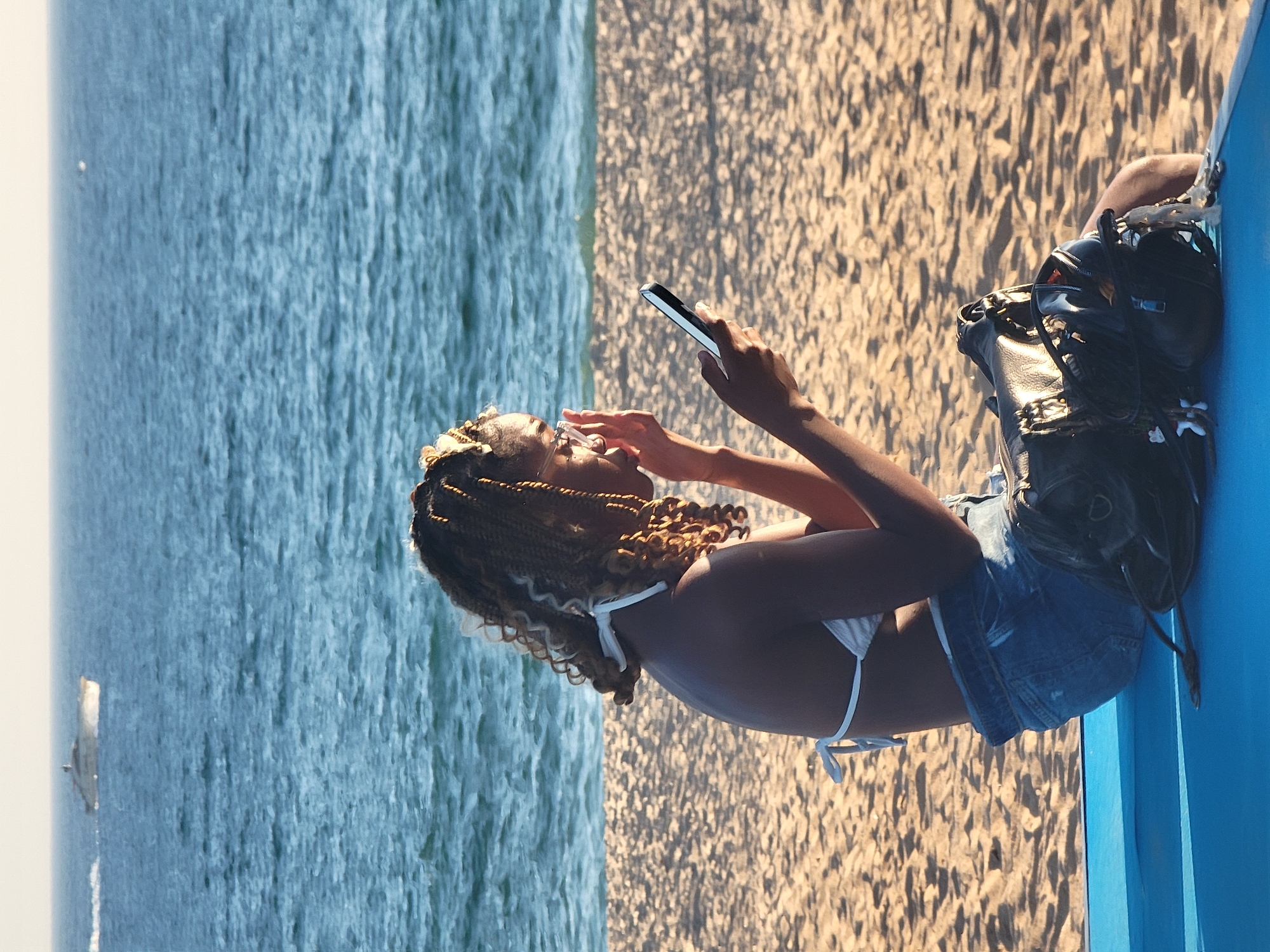 A woman with braided hair and glasses is sitting on a blue surface on a sandy beach in Quarteira, Portugal, looking at her phone. She is wearing a white bikini top and denim shorts. A black handbag is placed beside her. In the background, the ocean is visible with a boat in the distance. The sun is shining, casting shadows on the sand.