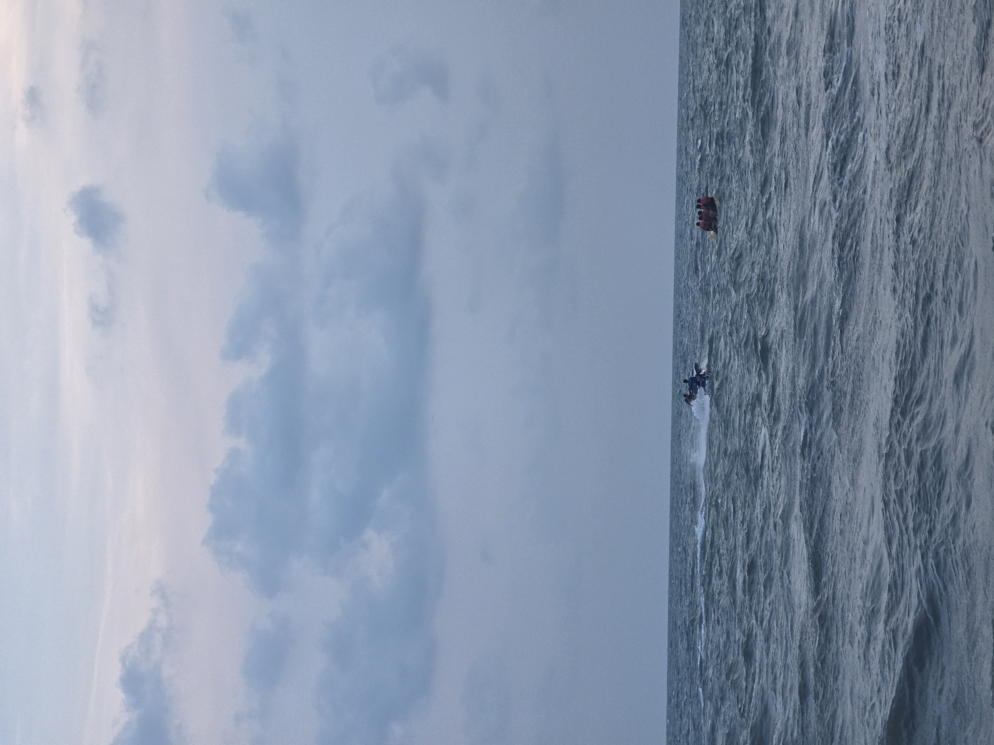 The scene is in Pattaya City, Thailand, and depicts a seascape with a partially cloudy sky. The sky occupies approximately half of the image, showing light blue hues and formations of clouds. The other half of the image displays a textured body of water with a motorboat traversing it. Behind the motorboat, a parasail is being pulled through the air, indicating the motorboat is engaged in parasailing activity. There may be people on the parasail, but they are difficult to distinguish due to their relative distance.