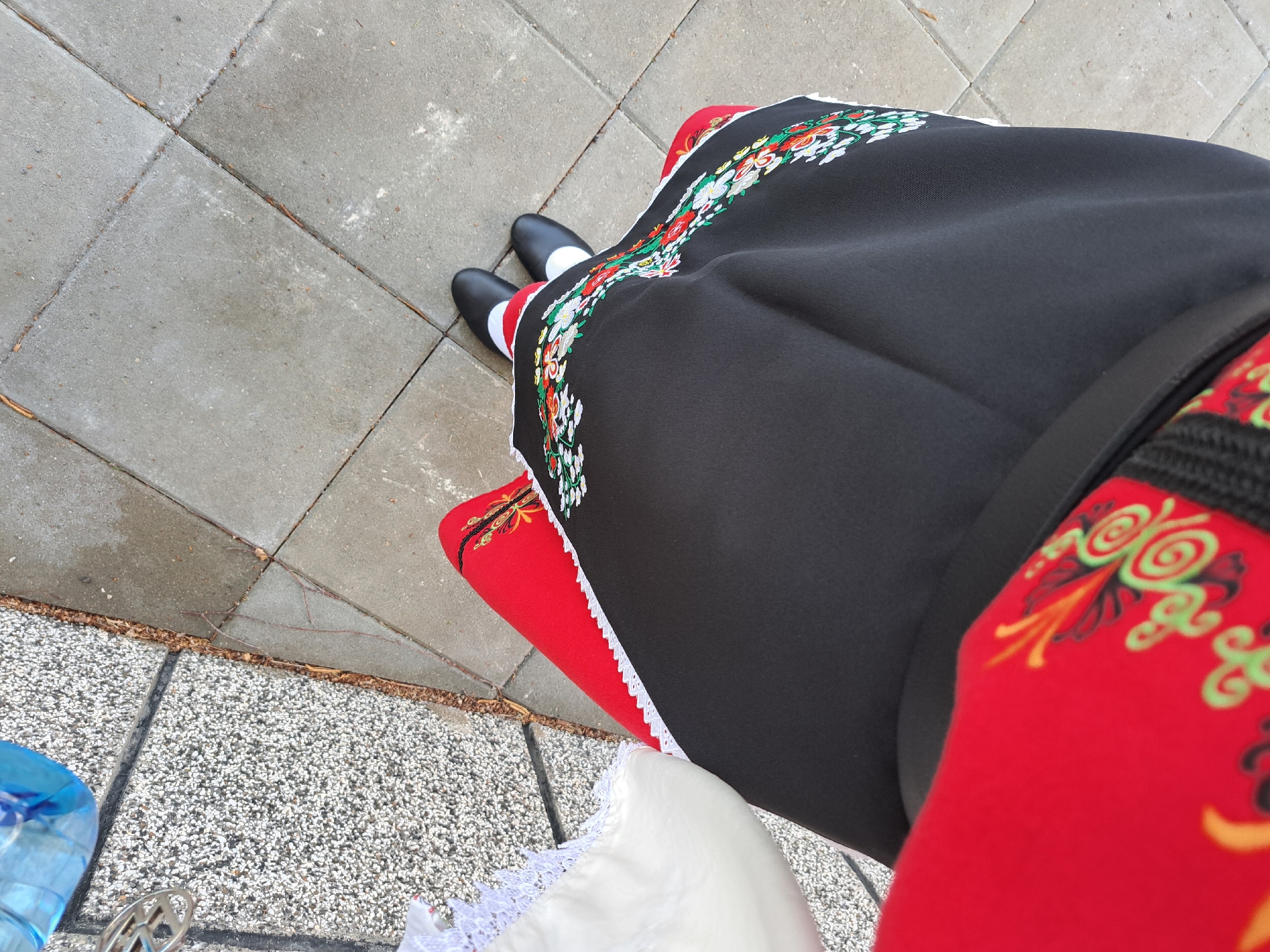 From an elevated, downward angle, the image depicts a person dressed in a traditional Bulgarian folk costume standing on a pavement in Lokorsko, Bulgaria. The person is wearing black shoes with white socks, a black skirt adorned with floral embroidery along the hem, a red underskirt, and a red apron with embroidered decorative elements. Portions of the pavement are visible in the corners of the frame, and what appears to be a water bottle in the lower left corner.