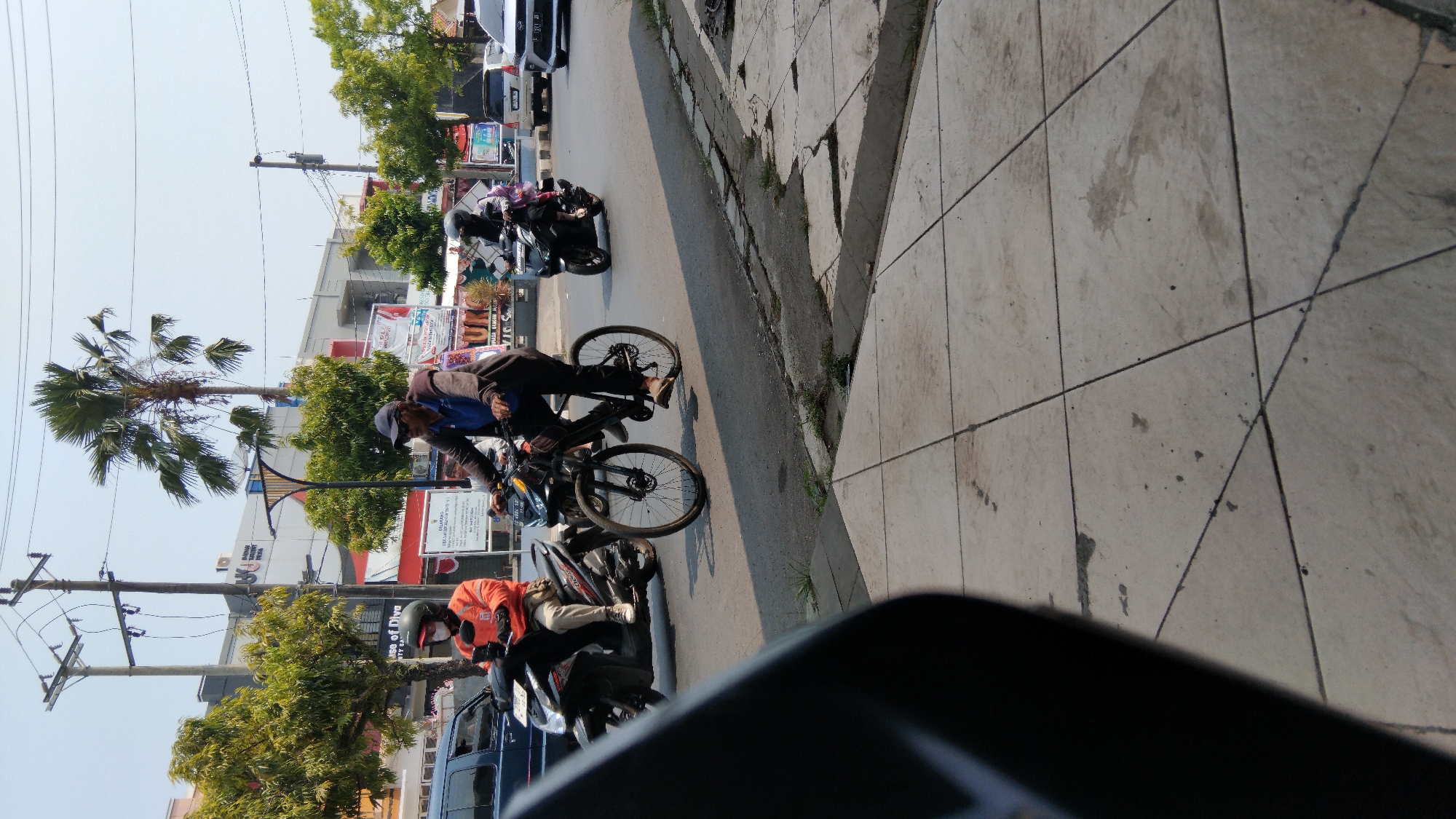 A person rides a bicycle on a paved road in Batang, Indonesia, on a sunny day. The cyclist, wearing a blue shirt, dark pants, a dark baseball cap, and sandals, is in the foreground. To the left of the cyclist, another person rides a scooter, wearing an orange jacket and a helmet. In the background, there are buildings, trees, and utility poles. Several other vehicles, including scooters and cars, are visible on the road. The scene is captured from a low angle, with a sidewalk in the foreground.