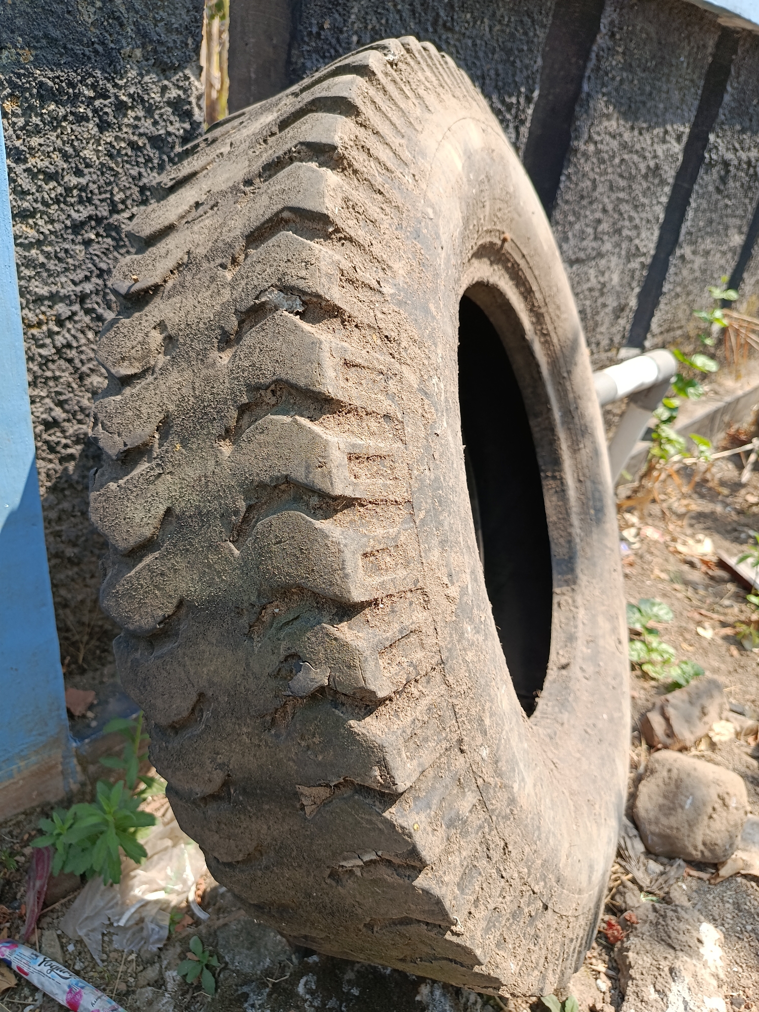 A close-up, slightly low-angle shot captures a large, worn, dirty tire leaning against a textured, dark gray concrete wall. The tire is the primary subject, positioned to the right of the frame, its treads deeply grooved and packed with dirt, indicating heavy use. The tire's sidewall is rough and shows signs of cracking and wear. To the left of the tire, a vertical segment of a bright blue painted surface is visible, possibly a post or pillar.
The background consists of the concrete wall, which is rough and uneven in texture, casting subtle shadows from an unseen light source. Beyond the wall, to the right, a lighter-colored, rough surface, possibly a cement walkway or structure, is visible, with sparse green vegetation growing around it. The ground in the foreground is a mix of dirt, small stones, dry leaves, and some scattered white plastic debris, suggesting an outdoor, possibly neglected, area. The lighting indicates a sunny day, with strong highlights and shadows. The overall impression is of an outdoor scene, possibly a storage area or a roadside, in Kemiri, Indonesia. No people or animals are present in the image.