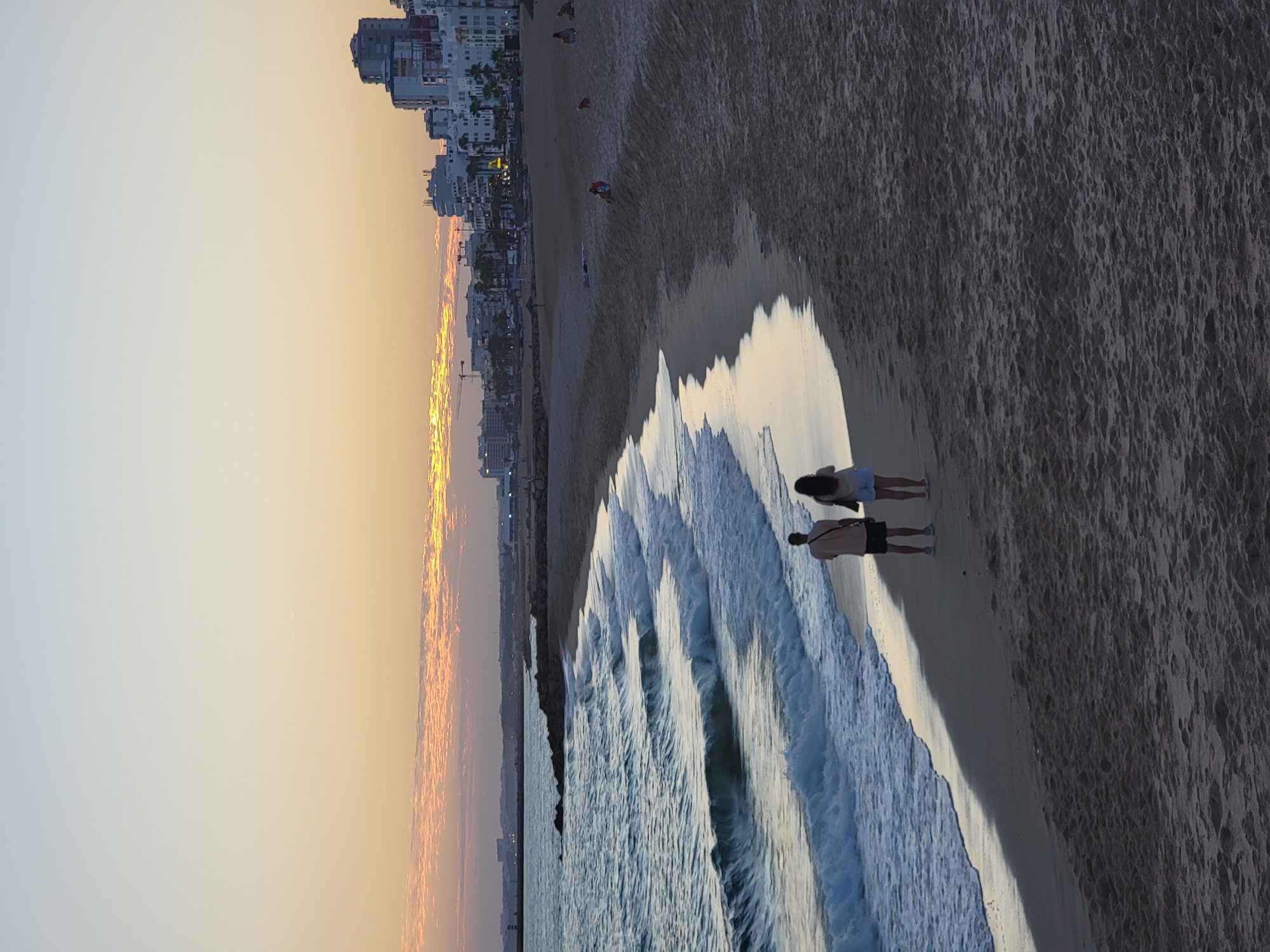 A vertical, eye-level, full shot captures the Algarve coastline in Quarteira, Portugal at dusk. The sky transitions from a light blue at the top to a warm orange and yellow hue near the horizon, with wispy clouds tinged pink from the setting sun. The ocean, with its white-capped waves, meets a sandy beach that curves into the distance. A line of rocky structures and a breakwater extend into the sea on the left side of the frame. In the background, the urban skyline of Quarteira is visible, with multi-story buildings and palm trees lining the coast. A few people are scattered on the beach, with two individuals, a man and a woman, walking together on the wet sand near the water's edge in the foreground. They are facing away from the camera and towards the ocean. The sand in the foreground is dark and textured, suggesting it is damp from the receding tide, and a long shadow is cast across it. The overall mood is tranquil and serene, characteristic of a peaceful evening by the sea.