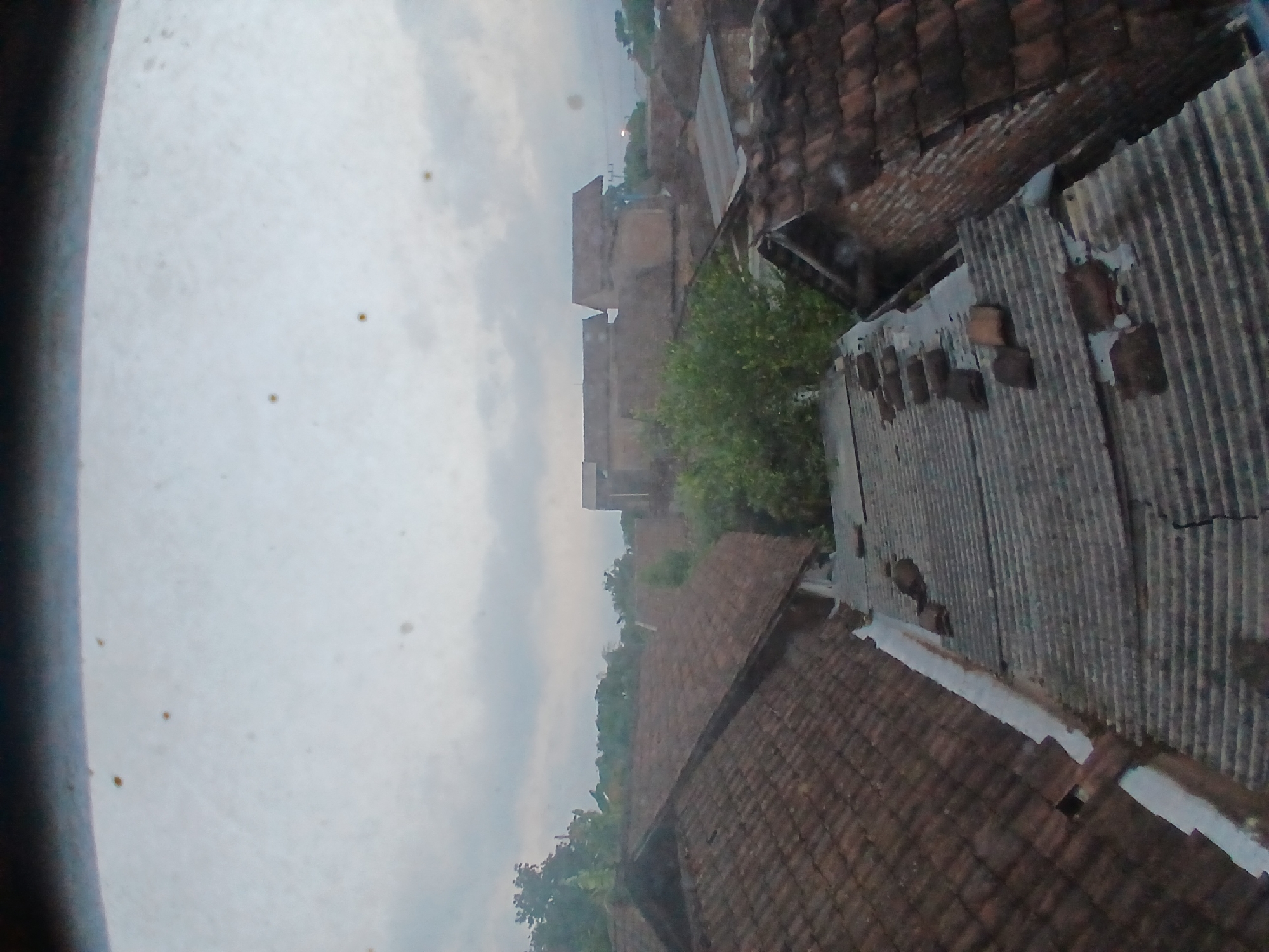 The image shows a view of rooftops in what appears to be a residential area in Tenggarang, Indonesia. The sky is overcast and a muted gray. The rooftops are primarily covered with terracotta tiles, with some corrugated metal roofing visible. In the foreground, there is a section of corrugated metal roof that appears weathered and has several loose, broken pieces of what might be tiles or bricks scattered on it. Adjacent to this, a pitched roof covered in terracotta tiles extends to the left. Further in the background, more rooftops and trees are visible, interspersed with what seem to be buildings. The overall impression is one of a rural or semi-urban environment, possibly during a cloudy day or early morning/late evening. There are no people or animals visible in the scene.
