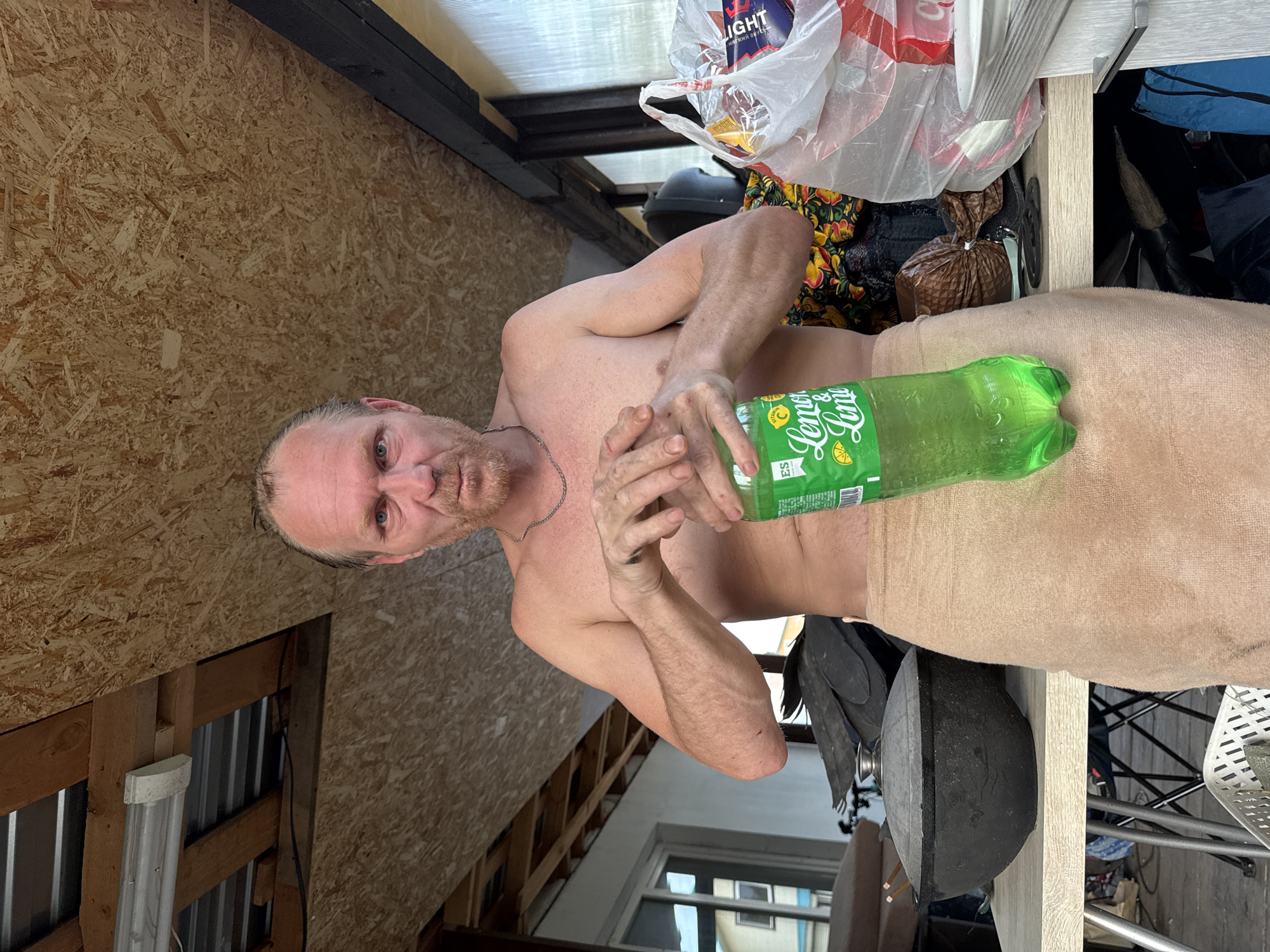 The image shows a shirtless man holding a green bottle of "Lemon & Lime" soda in front of his body, partially covered by a beige towel. He has light skin, a light beard, and appears to be frowning slightly. He is wearing a thin silver chain necklace.
Behind the man is a cluttered space. To his right, there is a table or shelf with various items on it, including a plastic shopping bag containing what seems to be a package labeled "LIGHT," and other indiscernible objects. Behind these items, there are what appear to be more containers or bags. Below the man is a dark object, which may be a cooking grill, and various other items, including what appears to be a metal chair and a brown package.
To the man's left is a wall made of plywood or OSB paneling. Below that, we can see the support structure of the wall and a glimpse of an exterior scene including windows, possibly indicating that the scene is indoors but near an exterior opening, such as a shed or partially enclosed porch.
Without specific metadata, there is no way to be sure of the location in Chaikovsky, Russia, but the scene may be an interior space possibly under construction or in disrepair, as indicated by the unfinished wall.