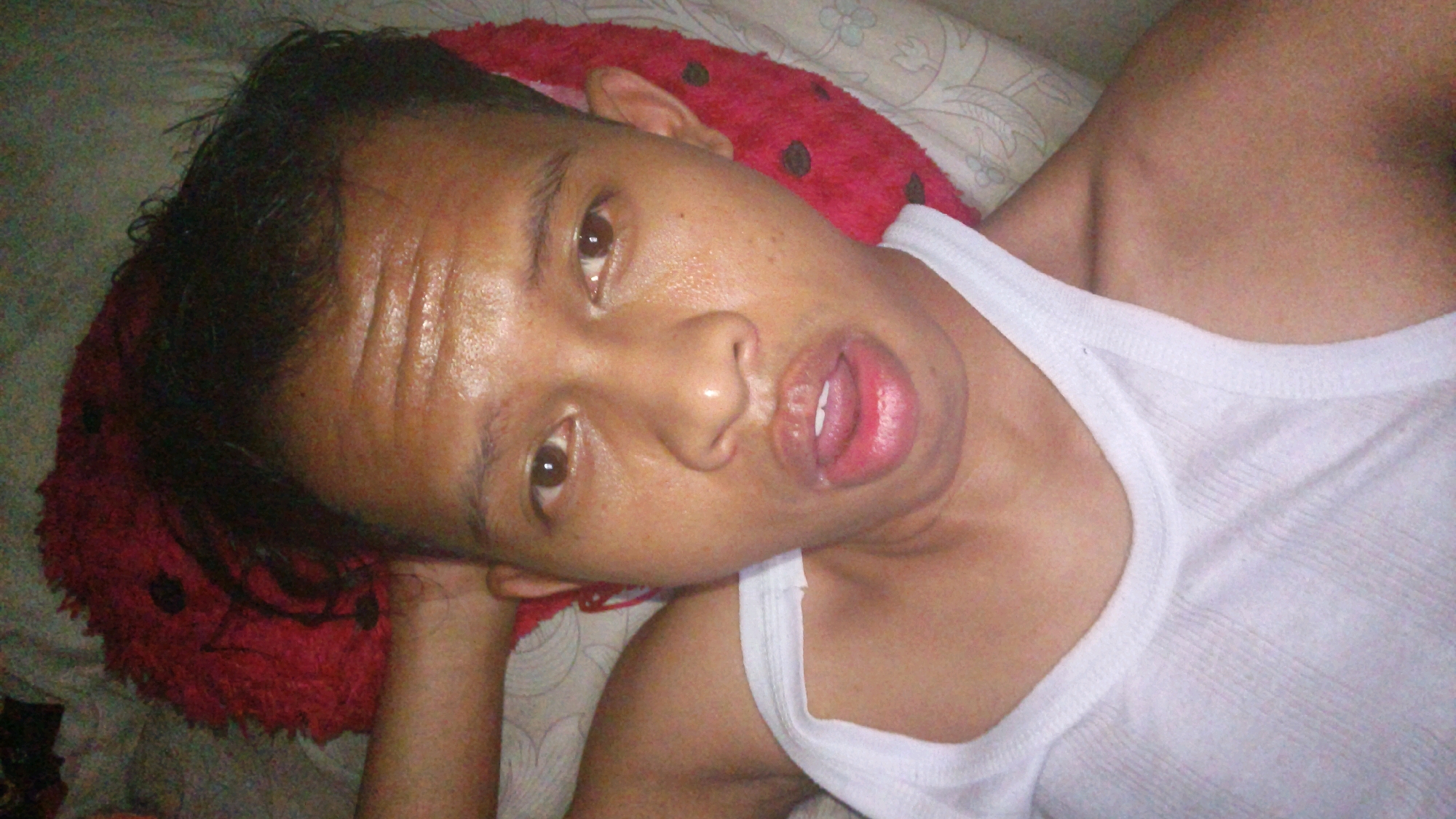 A person with dark hair and brown eyes, wearing a white tank top, is lying down with their head resting on a red pillow with black polka dots. The person's mouth is slightly open, revealing teeth. The background is a bed with a patterned sheet. The image appears to be a selfie taken in Pundungsari, Indonesia.