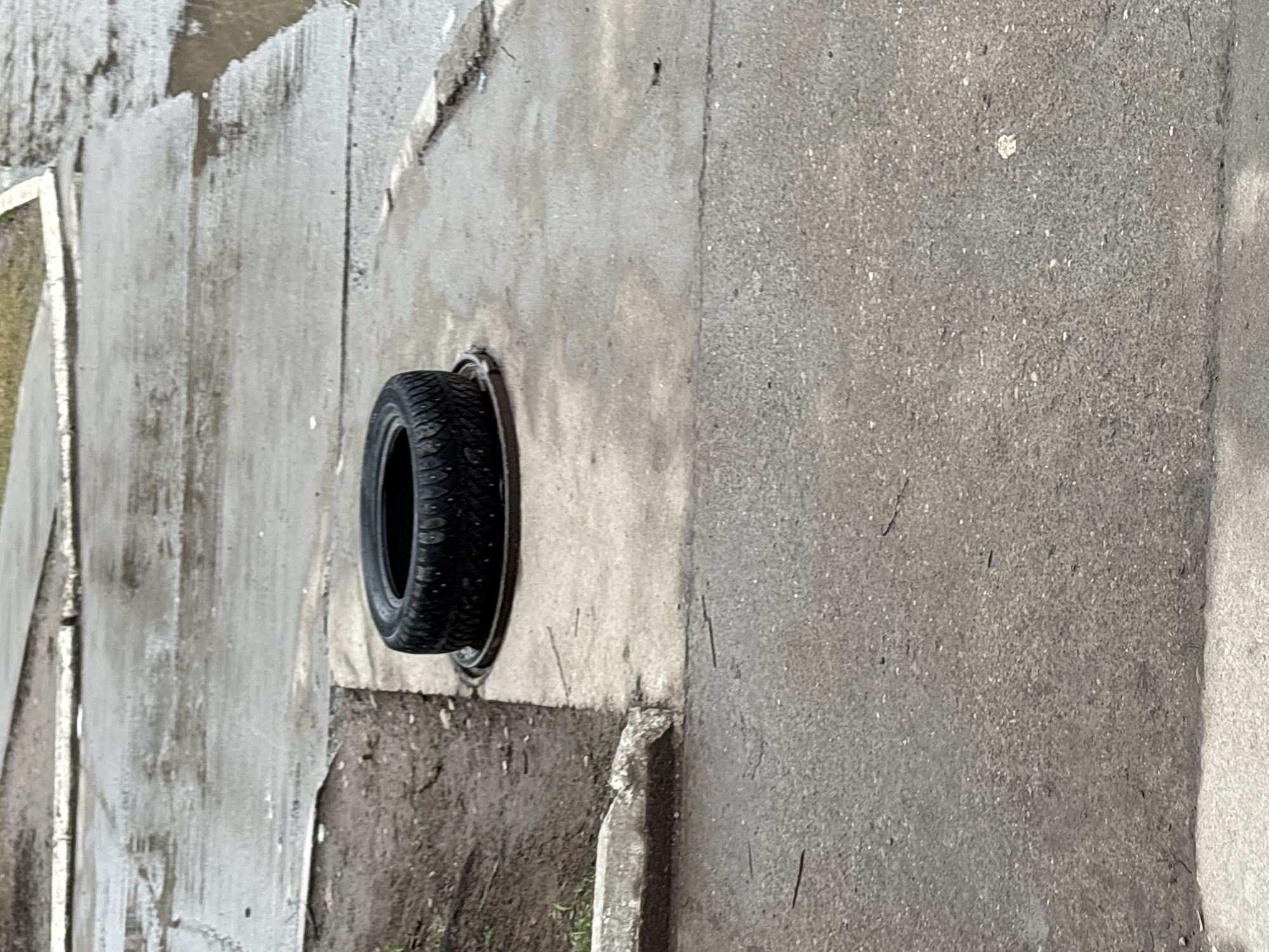 The image shows a black tire embedded in a concrete wall or structure. The surrounding area appears to be an outdoor setting, possibly a street or roadside. The concrete is weathered and marked. There is no indication of people or specific activities. The scene appears to be in a state of disrepair or neglect. There is no visible indication this is the city of Chaikovsky, Russia.
