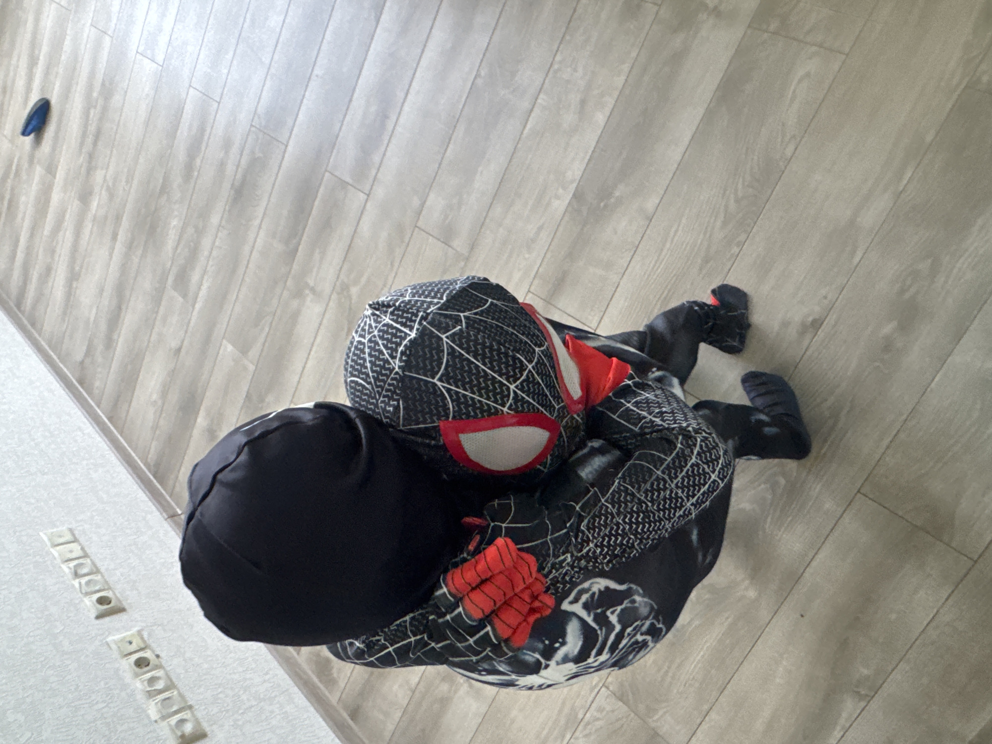 The image shows a person dressed in a Spider-Man costume, possibly a child due to the size of the costume and the setting. The costume is primarily black with white web patterns and red accents on the mask and gloves. The person is lying on a light-colored wooden floor, with their body curled up. One arm is tucked under their head, and the other is bent.
The setting appears to be indoors, on a floor made of light grey wood planks. To the left of the frame, there is a white wall with several electrical outlets visible. The lighting suggests it is daytime, but no specific time can be determined. There is no visible text in the image. The overall mood of the image is calm and relaxed, with the person appearing to be resting or playing on the floor. The dark blue object in the top left corner is likely a piece of clothing or a toy.